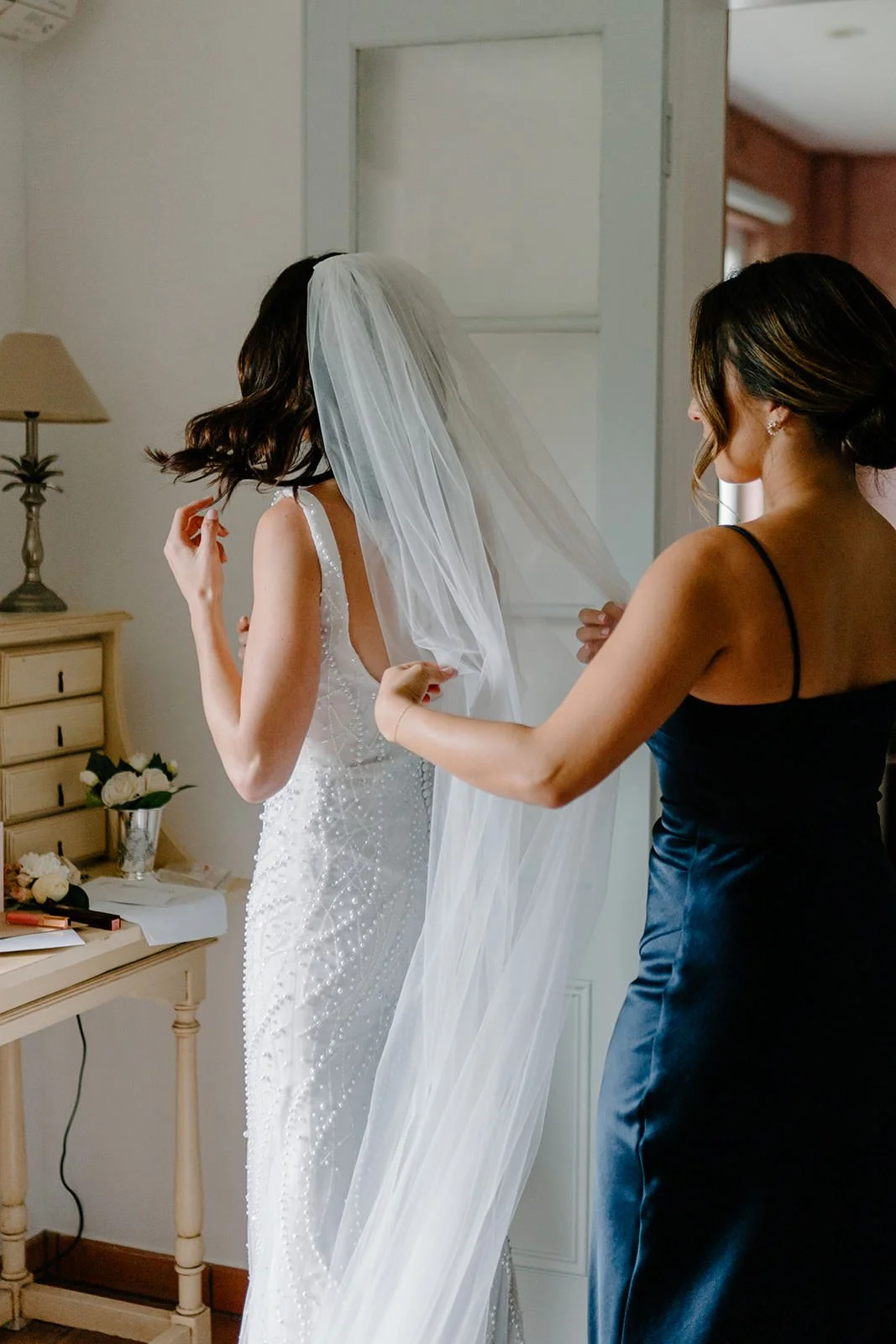 A bride wearing a white wedding dress with pearl embellishments is being assisted with her veil by a woman dressed in a black satin gown, in a room with a dresser and floral arrangements.