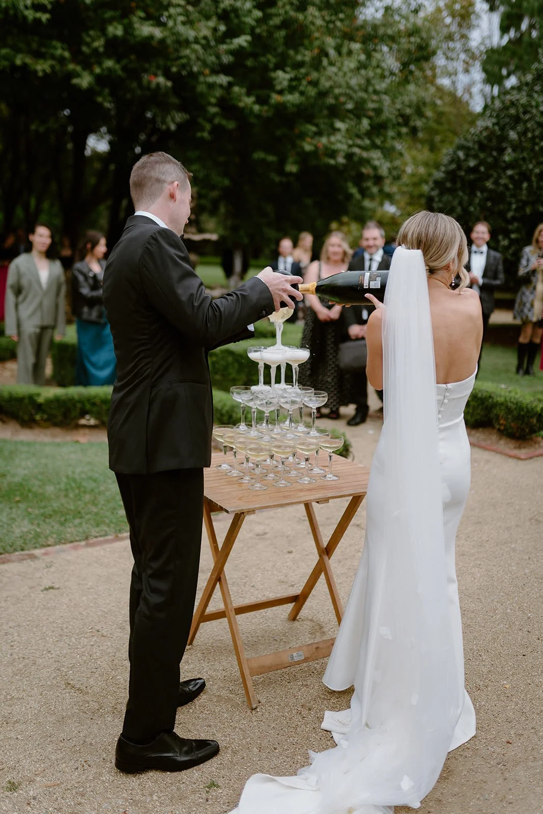 A wedding toast with a champagne tower in an outdoor garden, featuring a groom pouring champagne onto a bride's head, surrounded by guests in formal attire.