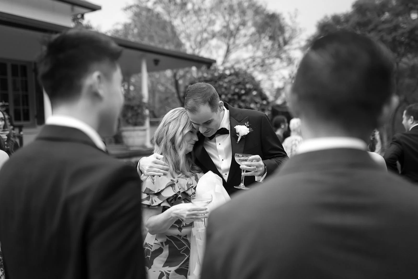 A black and white photo of a wedding reception outdoors, with a groom in a tuxedo and a woman sharing an emotional moment, surrounded by other guests in formal attire.