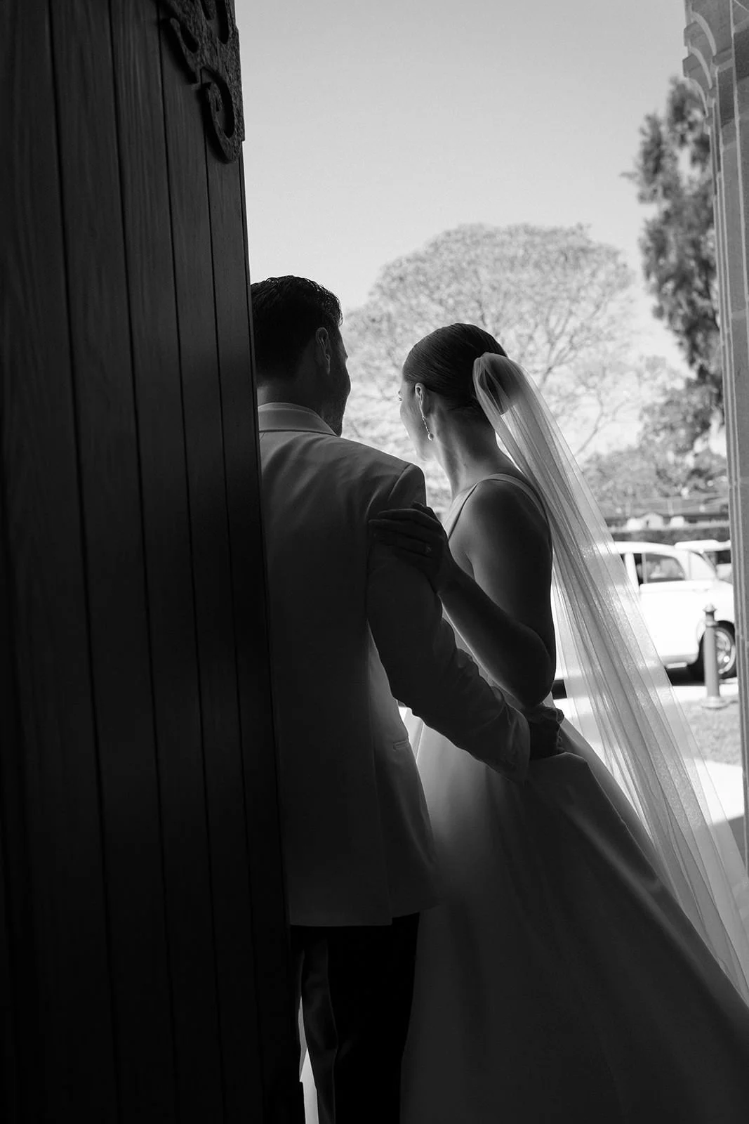 Black and white photo of a bride and groom sharing an intimate moment in a doorway, with trees and cars visible outside.