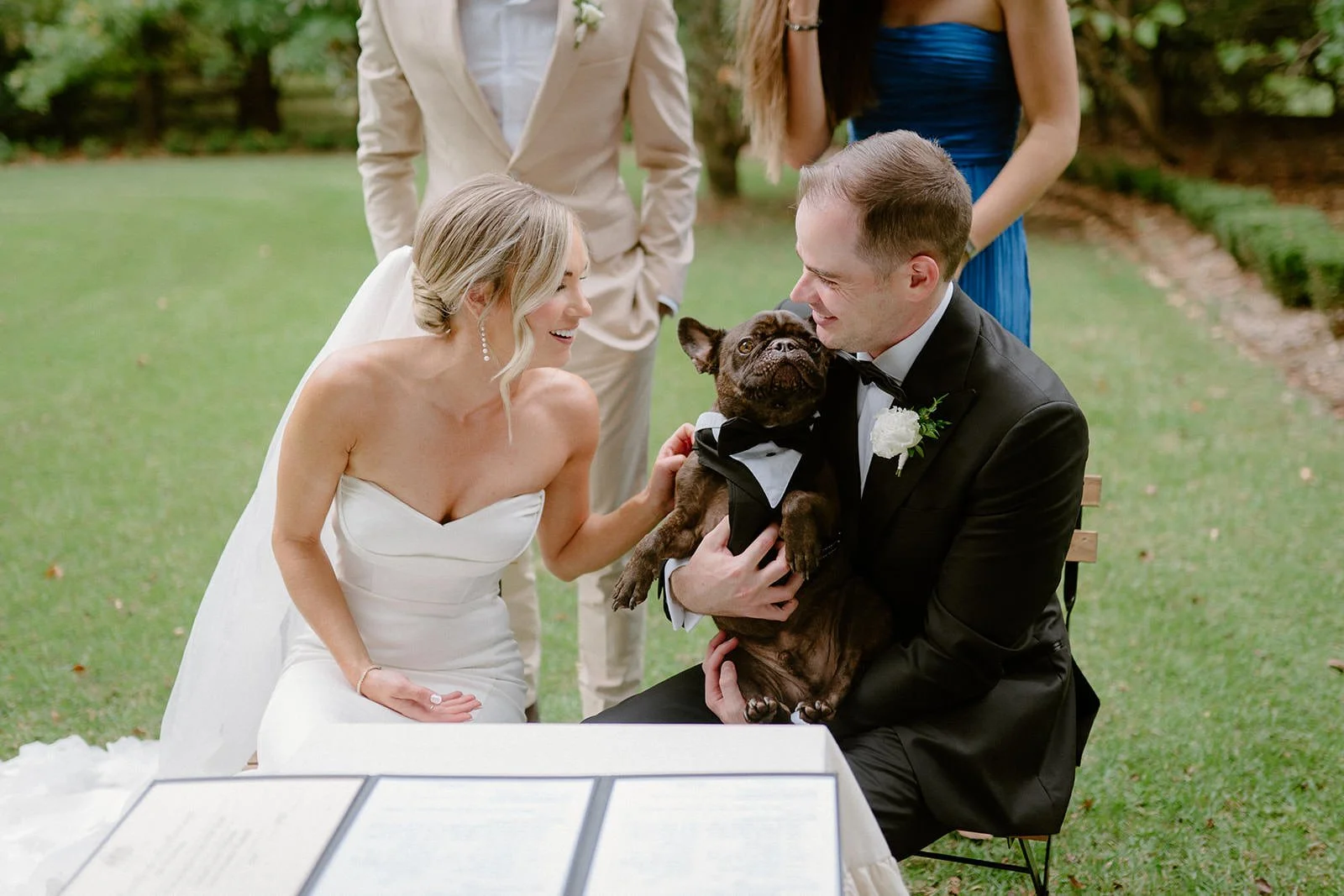 A bride and groom at their wedding, smiling and looking at each other, with a French bulldog dressed in a tuxedo between them. Two other people are standing behind, outdoors on a grassy lawn.