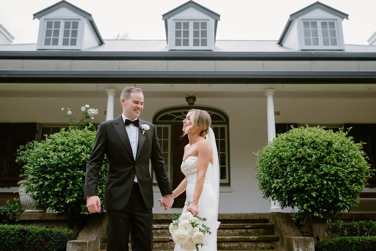 A bride and groom holding hands and smiling at each other in front of a white house with stairs and bushes.