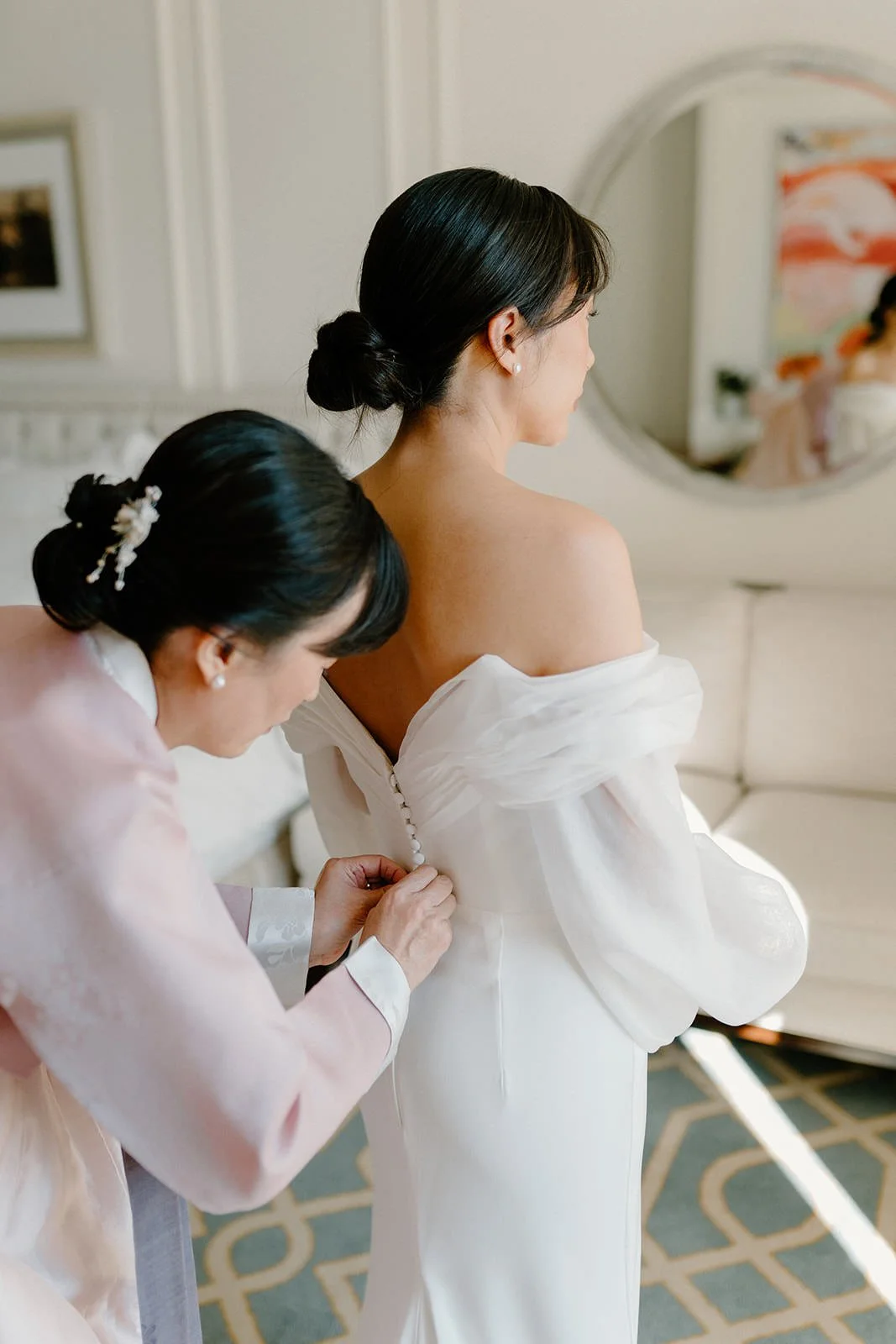 A woman in a white wedding dress is helping another woman, dressed in silk pink, button up her dress. The bride is facing away and the other woman is leaning forward, working on the buttons.