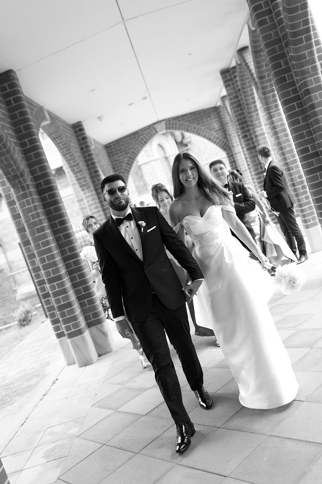 Black and white photograph of a bride and groom walking together, surrounded by wedding guests, under a brick archway with curtains, during a wedding celebration.