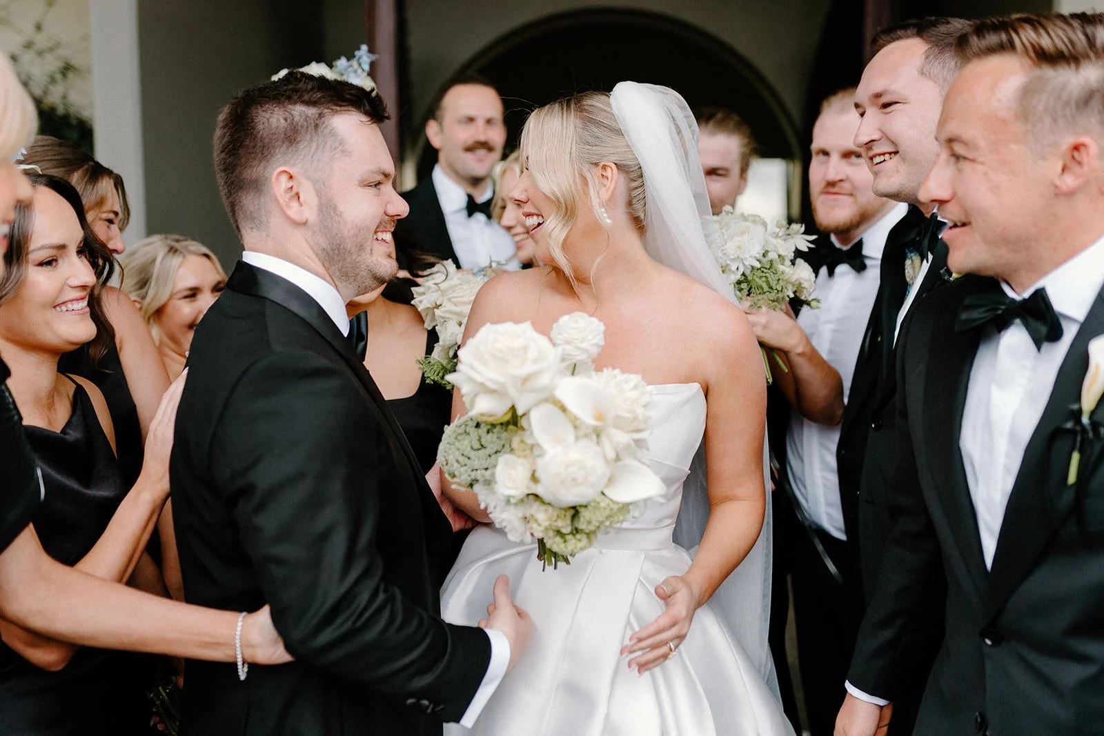 Bride and groom smiling and holding hands surrounded by friends in formal attire at wedding reception.