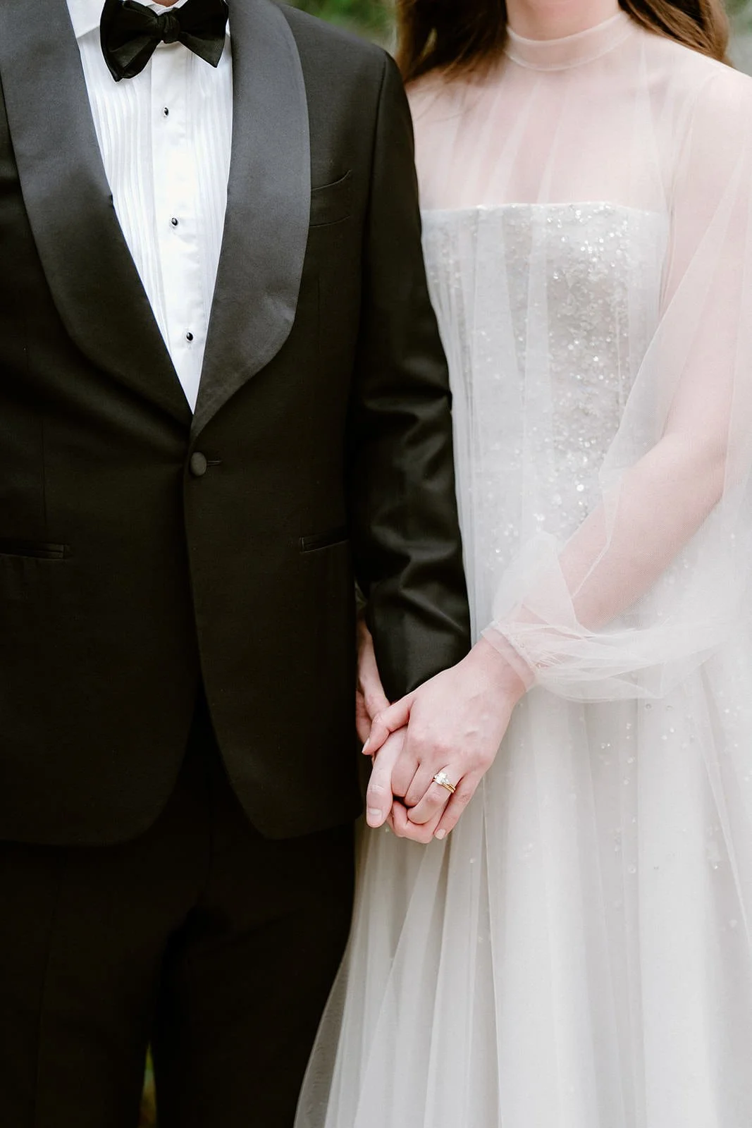 A bride and groom holding hands, with the bride wearing a white wedding dress and a wedding ring, and the groom wearing a black tuxedo.