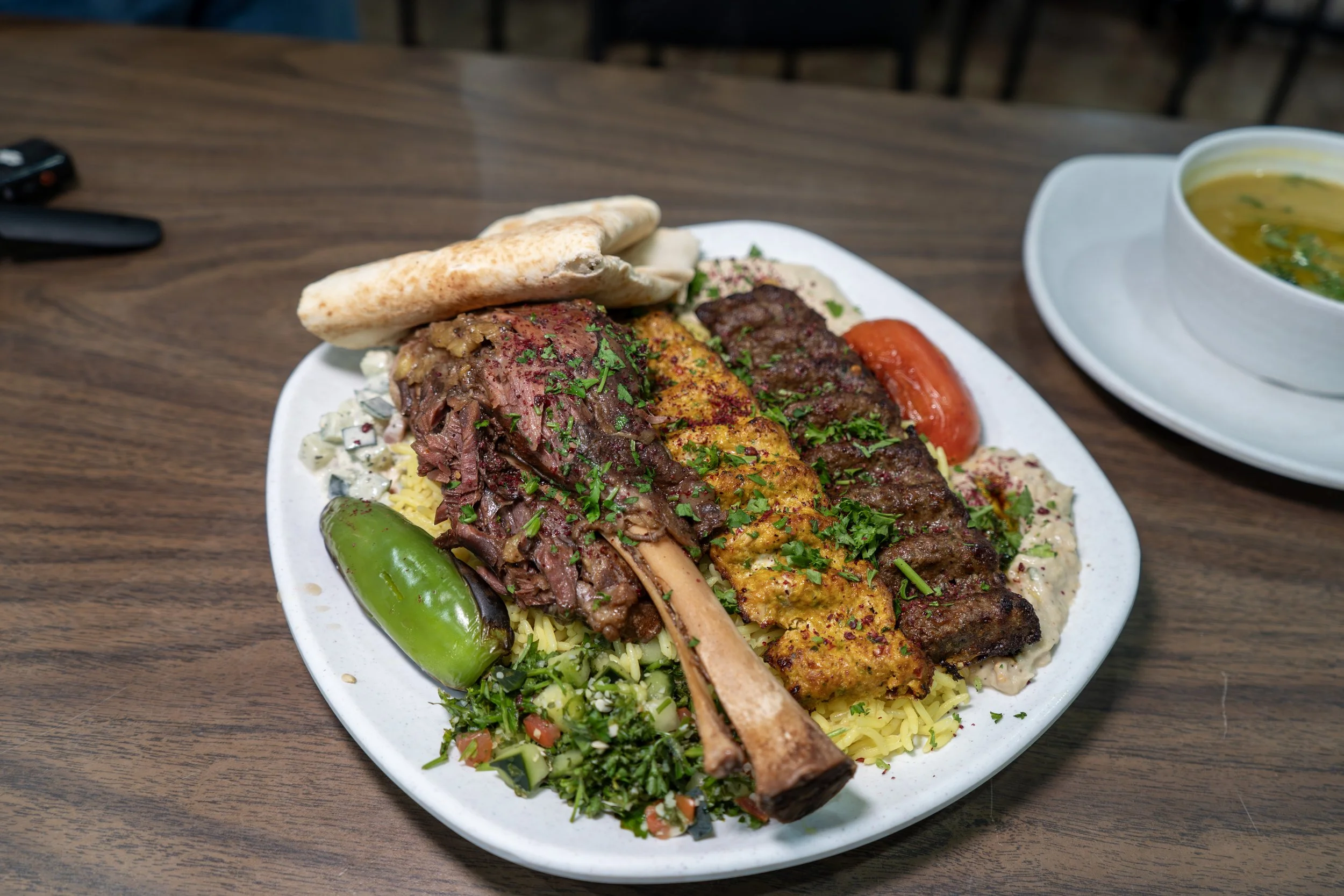 Image of a lamb shank, chicken and beef kabob on top of a bed of rice, grilled vegetables, tabbouleh, tahini salad, baba ghanoush, and hummus with a pita on top.