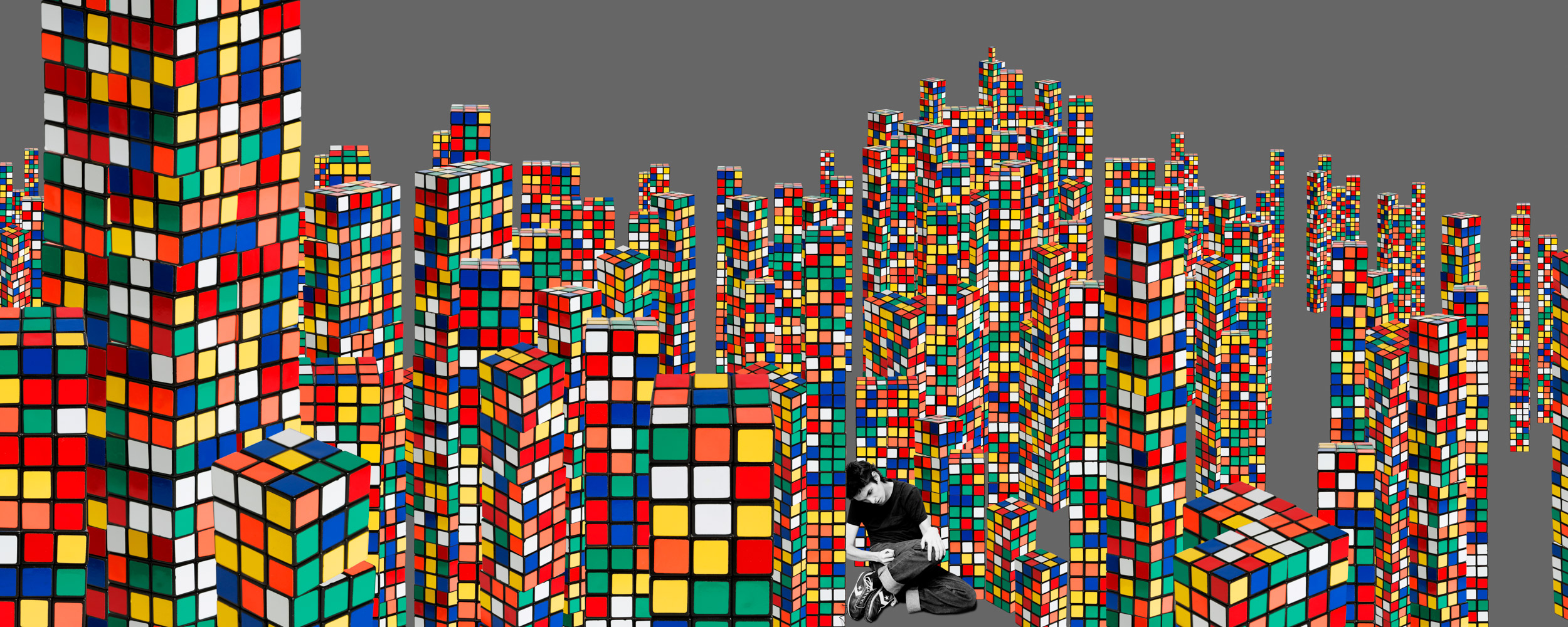 A person sitting amidst a cityscape made of giant, colorful Rubik's cubes on a gray background.