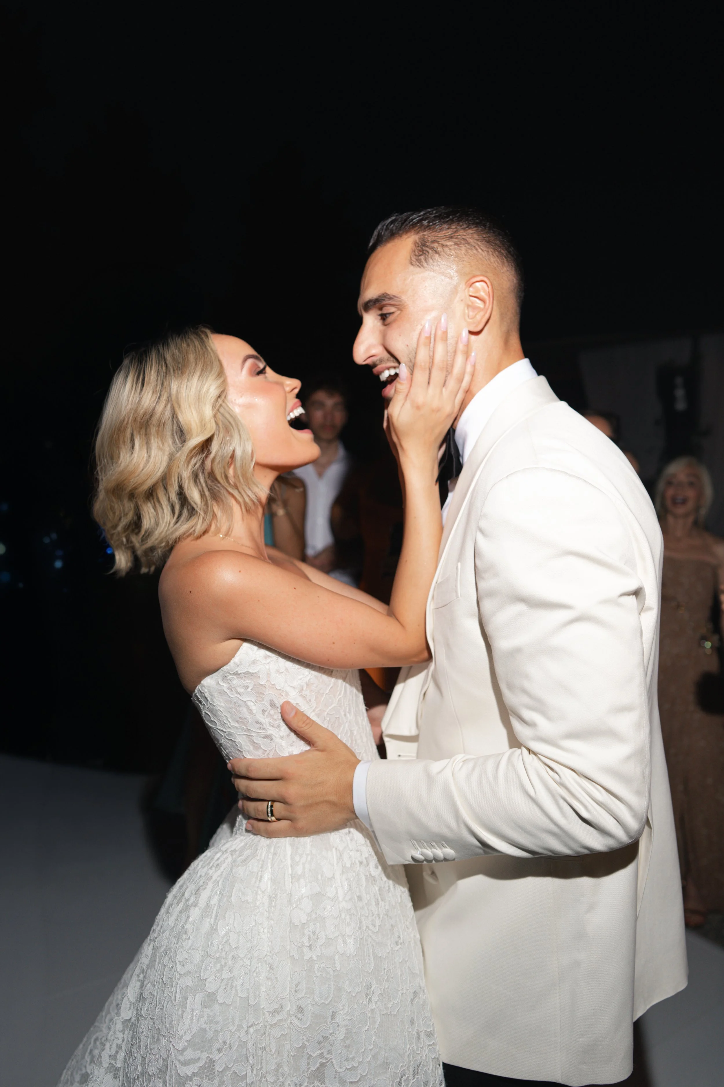 A bride and groom are dancing and smiling at their wedding reception, with the bride touching the groom's face and the groom looking at her affectionately.