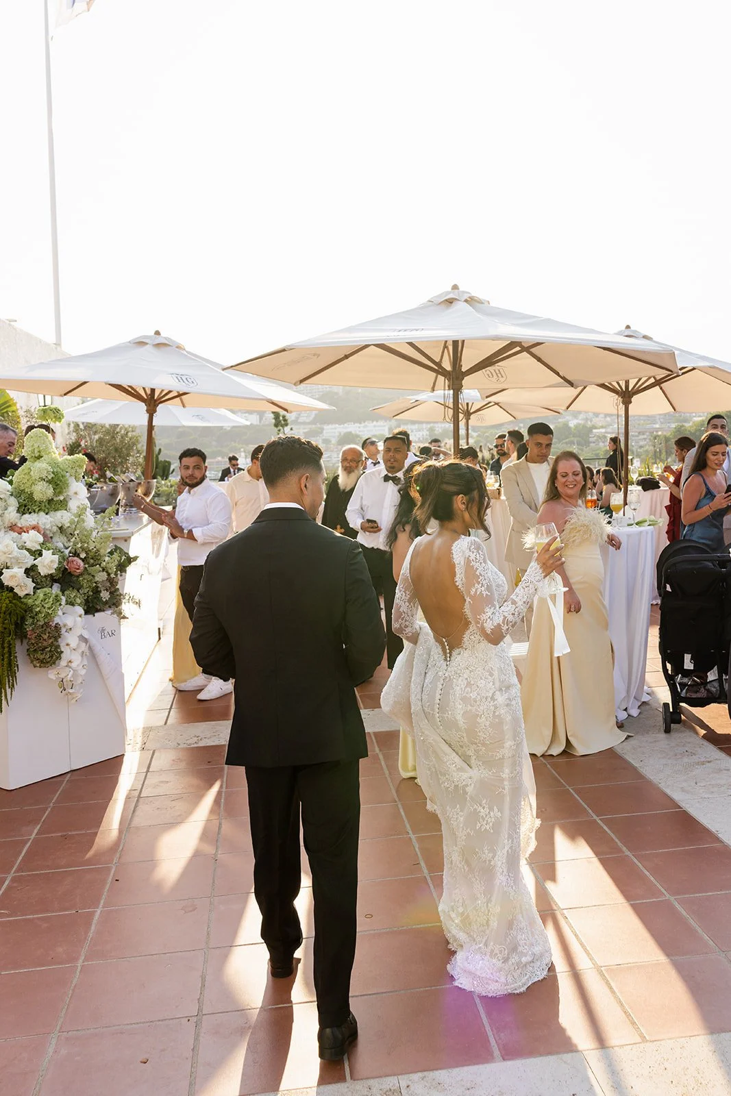 Bride and groom at an outdoor wedding reception, with guests mingling under large white umbrellas and scenic background.