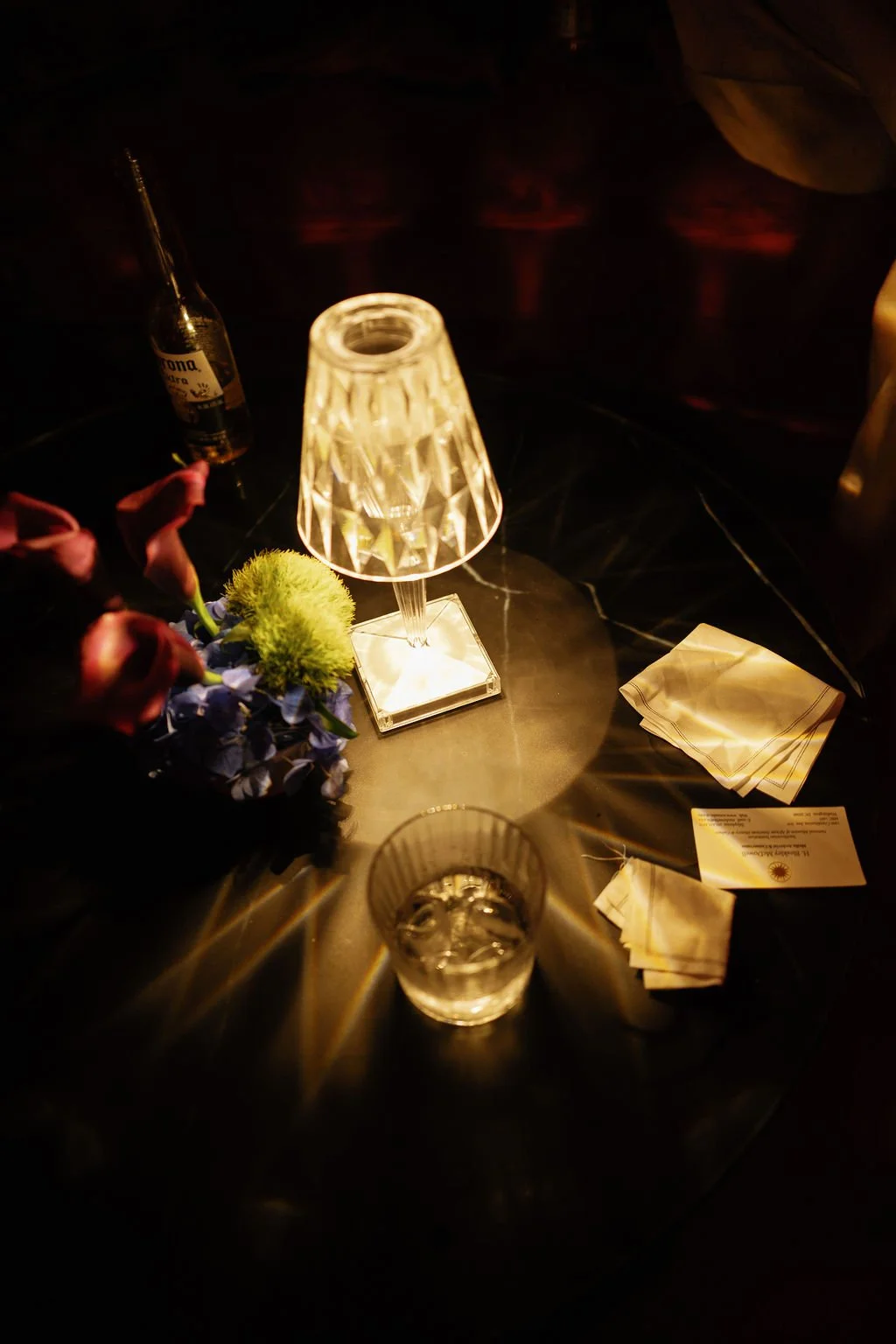 A dimly lit table with a lamp, a glass of water, a bottle of Corona beer, a small bouquet of flowers, and menus or napkins.