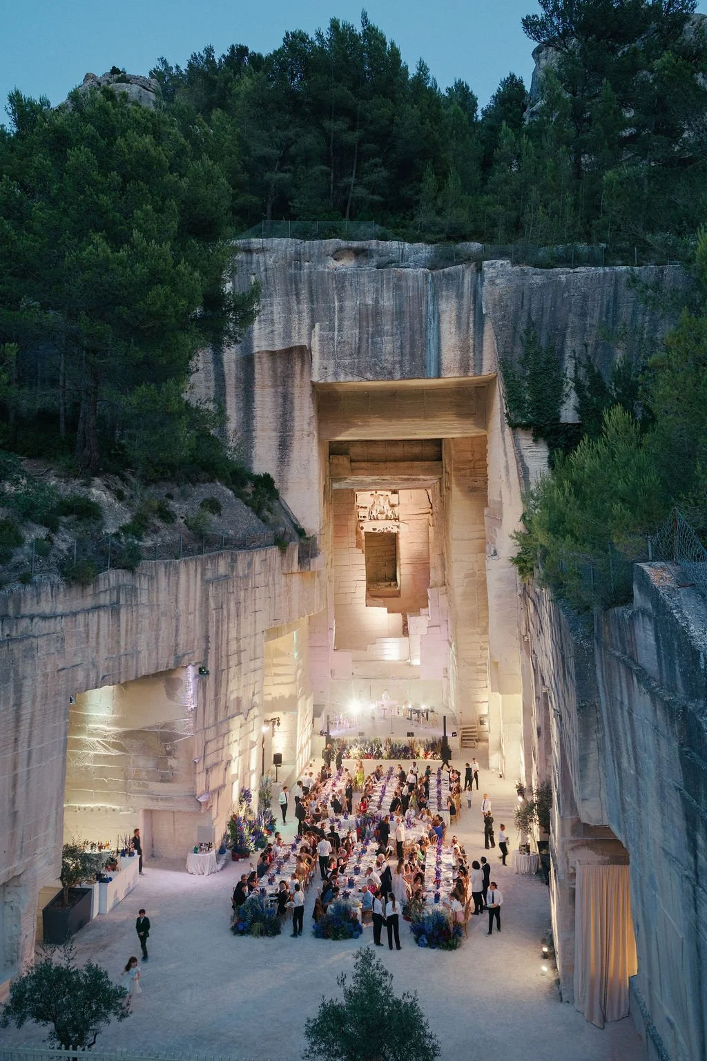 A large outdoor concert venue built into a rock cliff with a stage and a crowd of people dining at banquet tables under a covered area, surrounded by trees and mountainous landscape.
