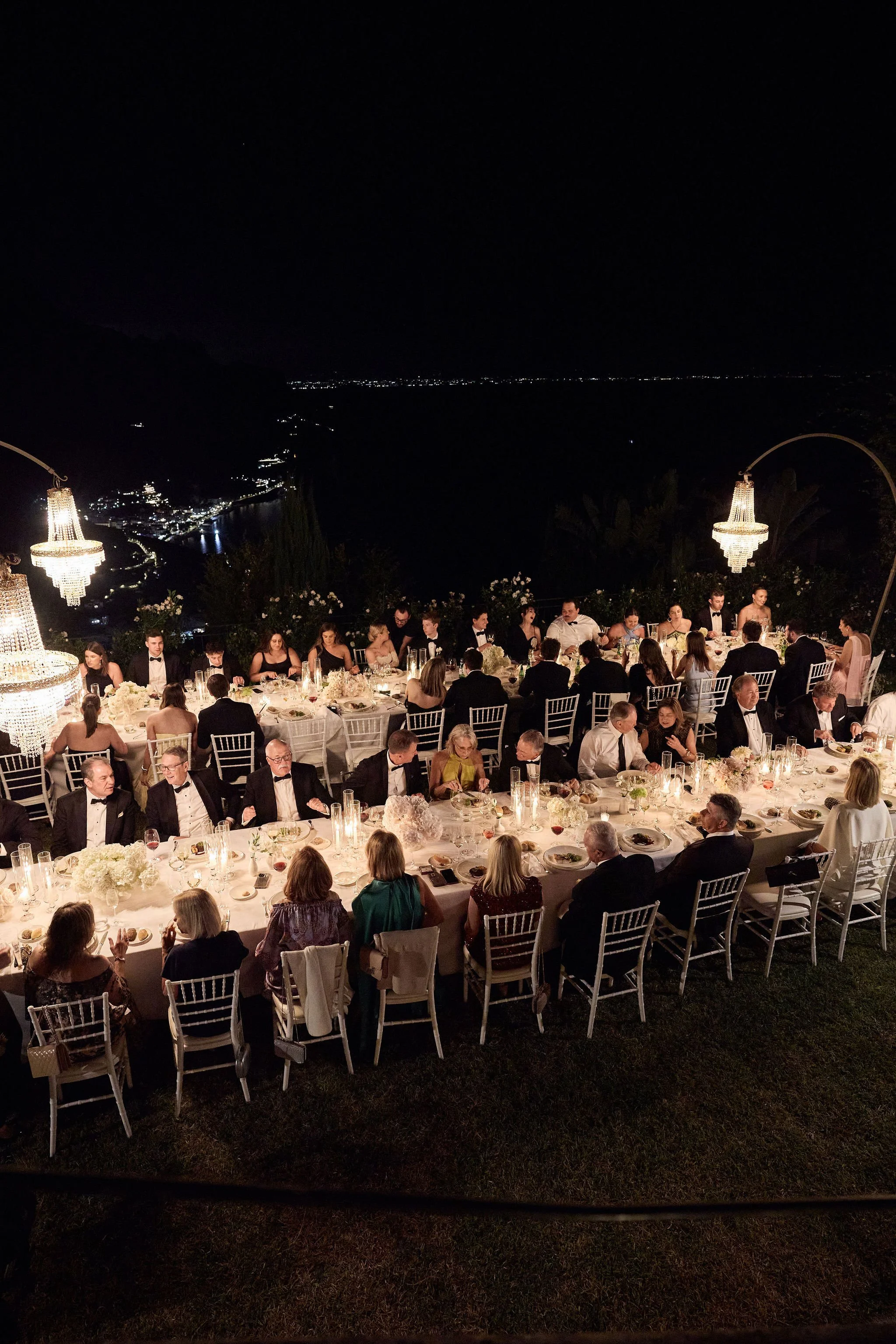 A nighttime outdoor banquet with many guests seated at long tables decorated with white flowers, served under chandeliers, with a lake and city lights visible in the background.