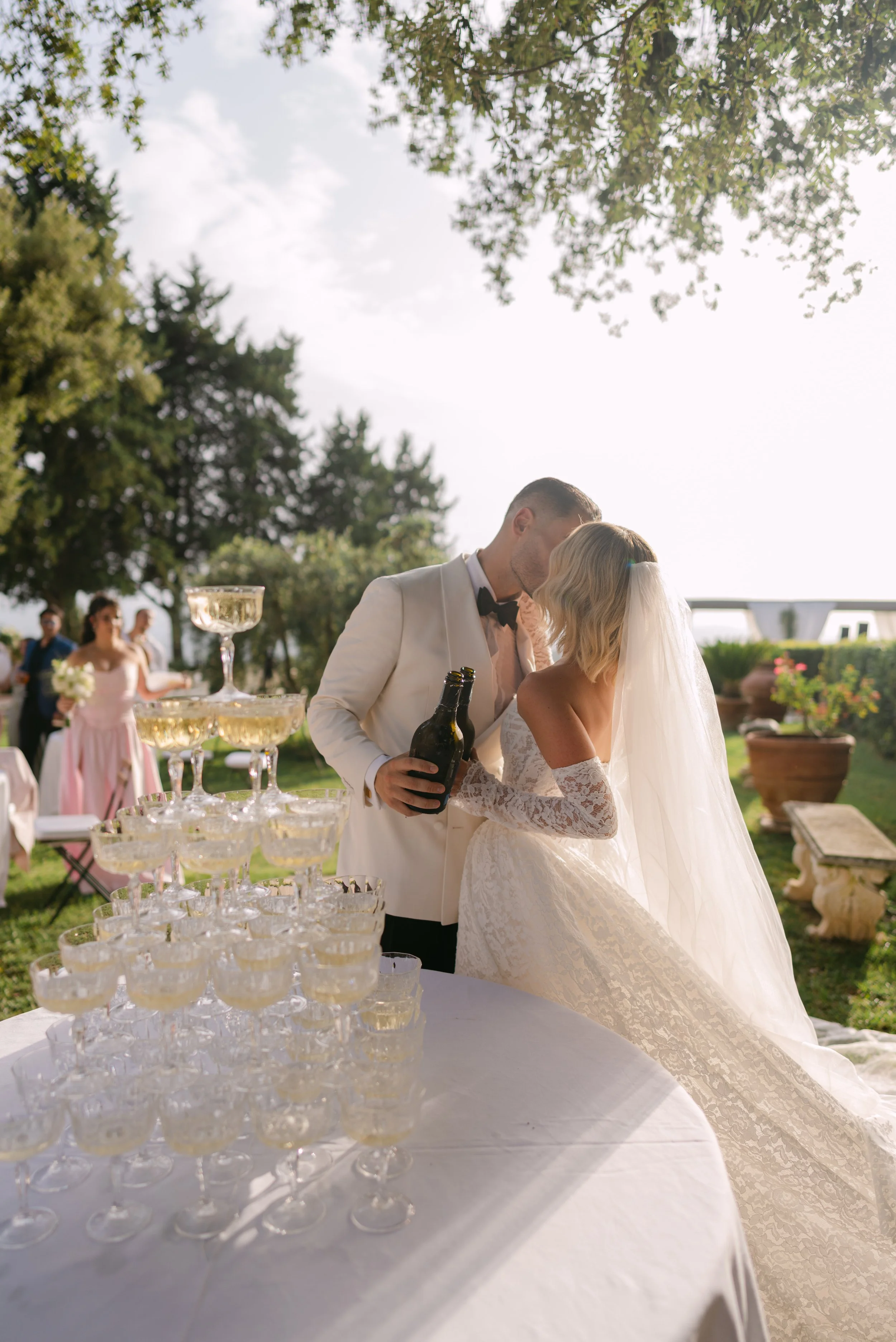 A bride and groom kissing at their wedding reception outdoors. A table with champagne glasses in the foreground. Bridesmaids and guests in the background, trees, and a bright sky.