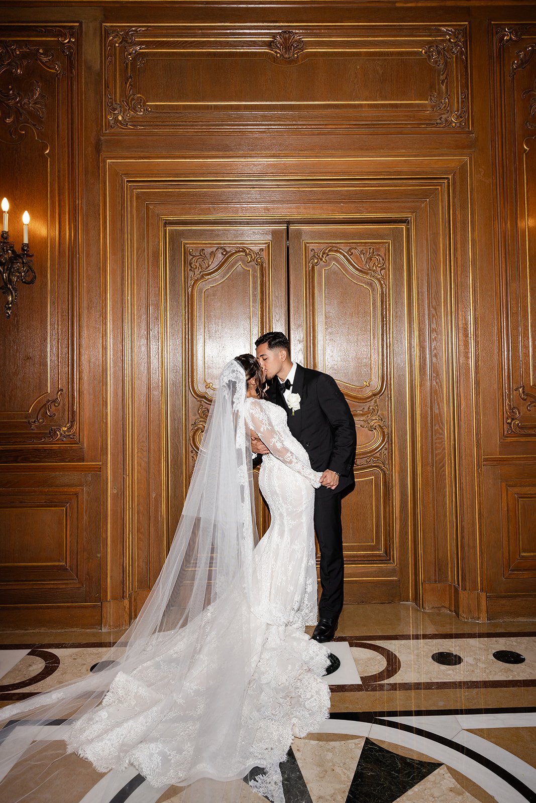 A bride in a white lace wedding dress with a long veil and a groom in a black tuxedo sharing a kiss in front of ornate wooden doors in an elegant hall.