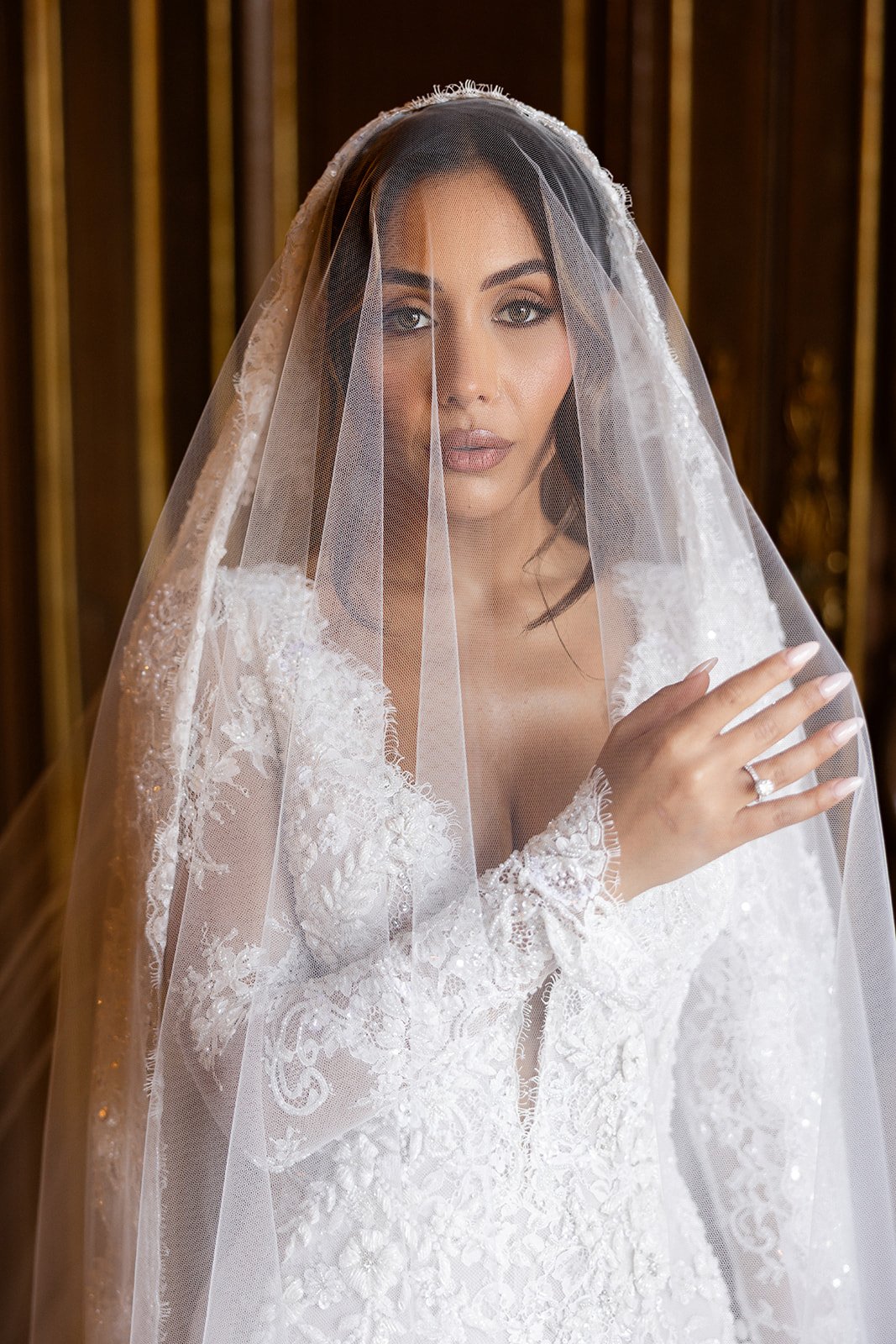 A woman in a wedding dress and veil looking at the camera, holding her hand near her chest with a ring visible.