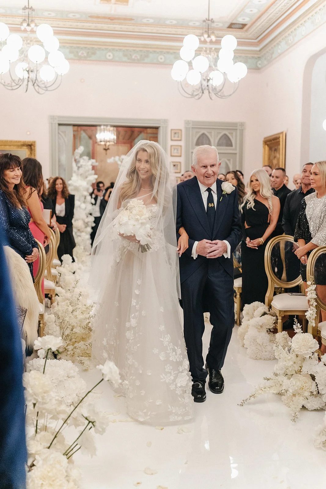A bride walking down the aisle with her arm linked with an older man during a wedding ceremony in an elegant decorated hall, surrounded by guests. The bride wears a white wedding gown with floral patterns and a veil, and is holding a bouquet of white