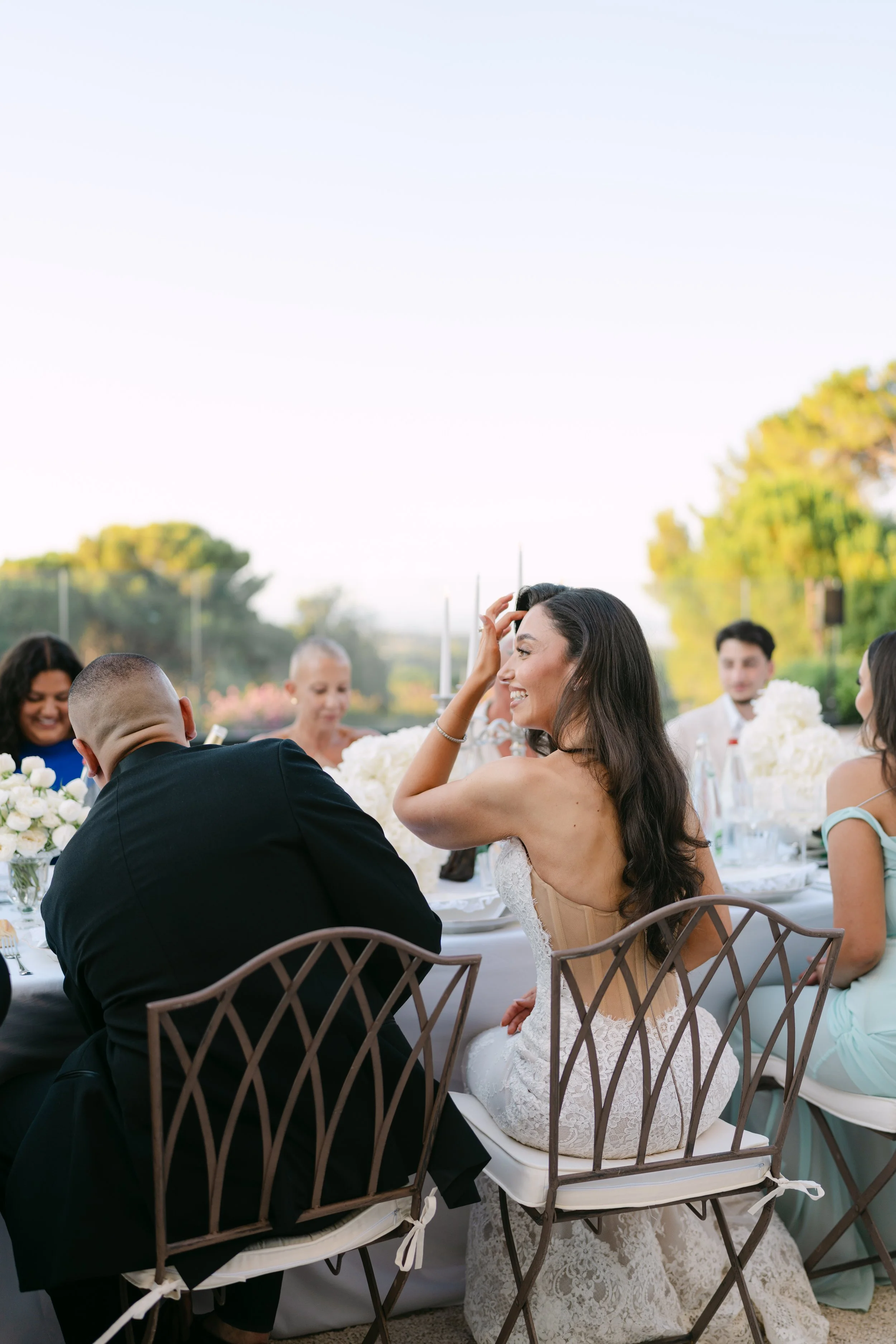 A bride laughing and smiling at a wedding reception, seated at a table with guests during an outdoor ceremony. The table is decorated with white flowers, and the guests are dressed formally. The background shows trees and clear sky.