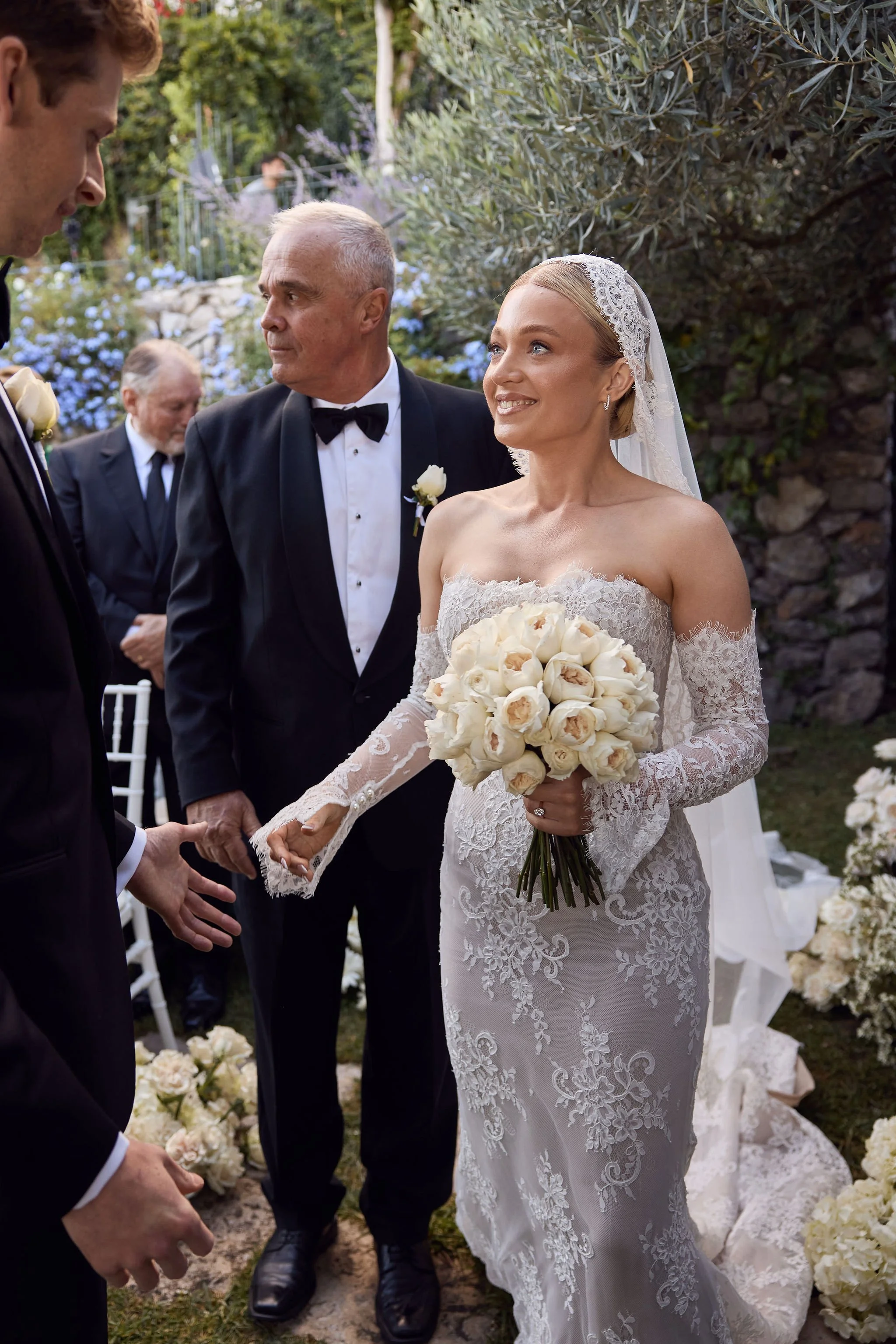 A bride in a lace wedding dress holding a bouquet of white roses, smiling at the groom during an outdoor wedding ceremony. Groomsmen and an officiant stand nearby in formal attire, with greenery and flowers in the background.