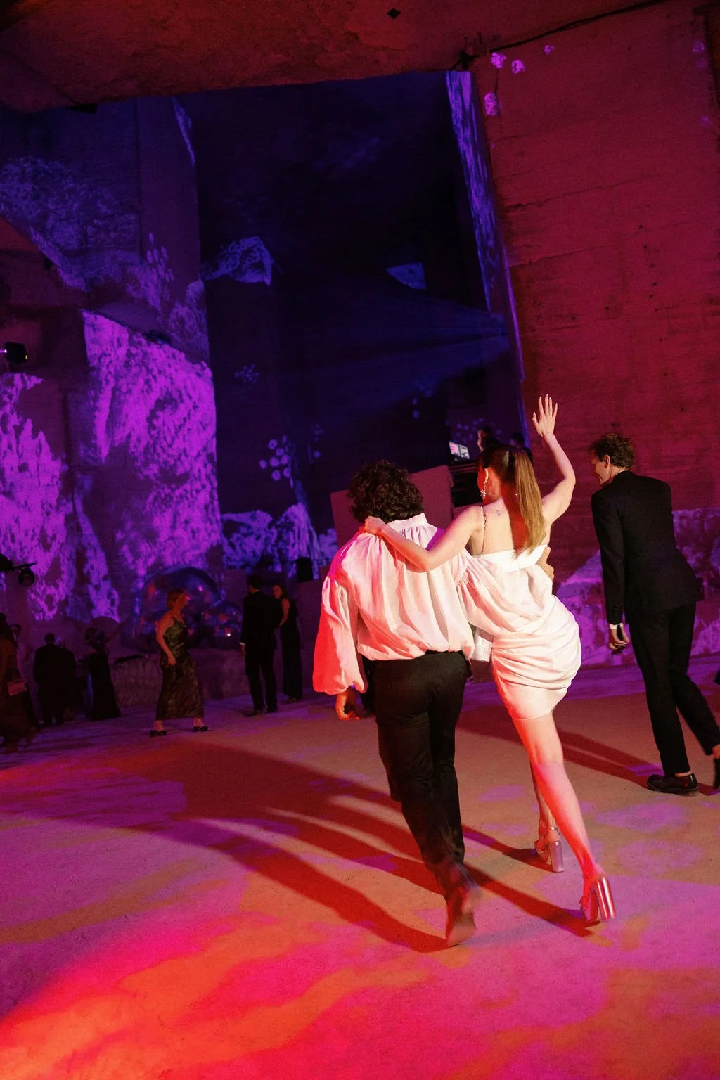 People dancing at an indoor event with colorful lighting and a large textured wall in the background.