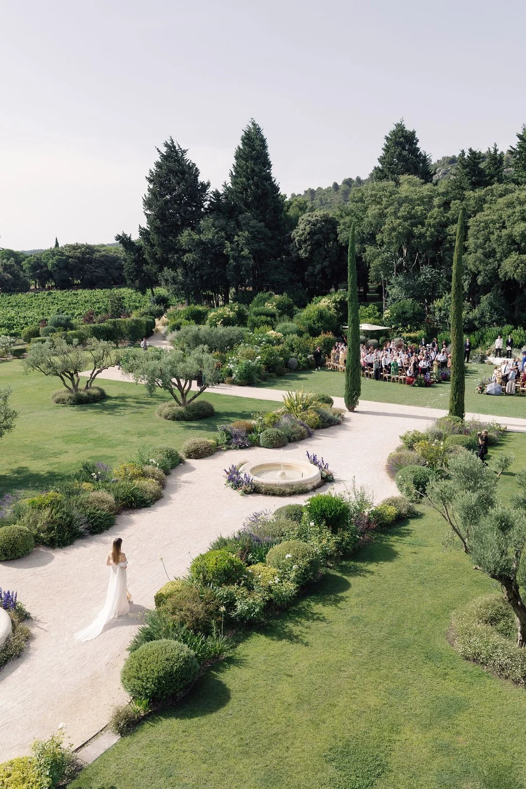An outdoor garden scene with a bride in a white wedding dress walking along a gravel pathway lined with manicured bushes and small trees. In the background, there's a group of people gathered for a wedding reception, seated at long tables under an um