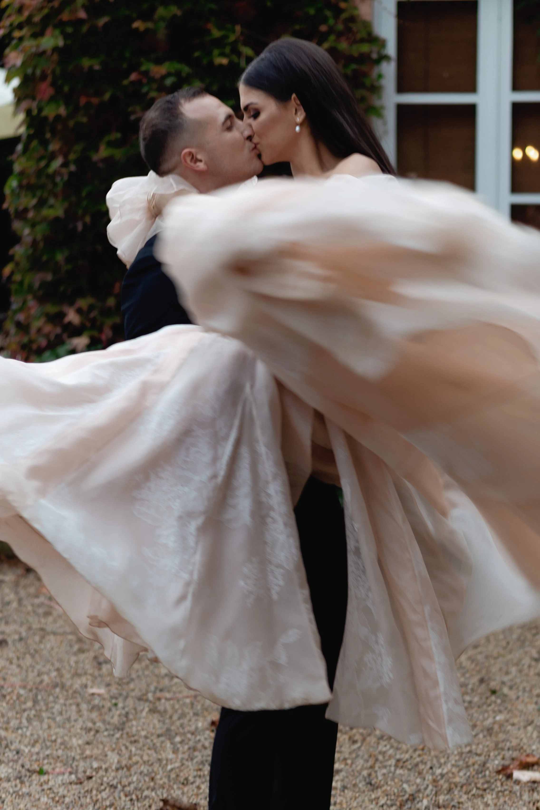 A couple sharing a kiss, with the man carrying the woman in his arms, outdoors near a building with windows.