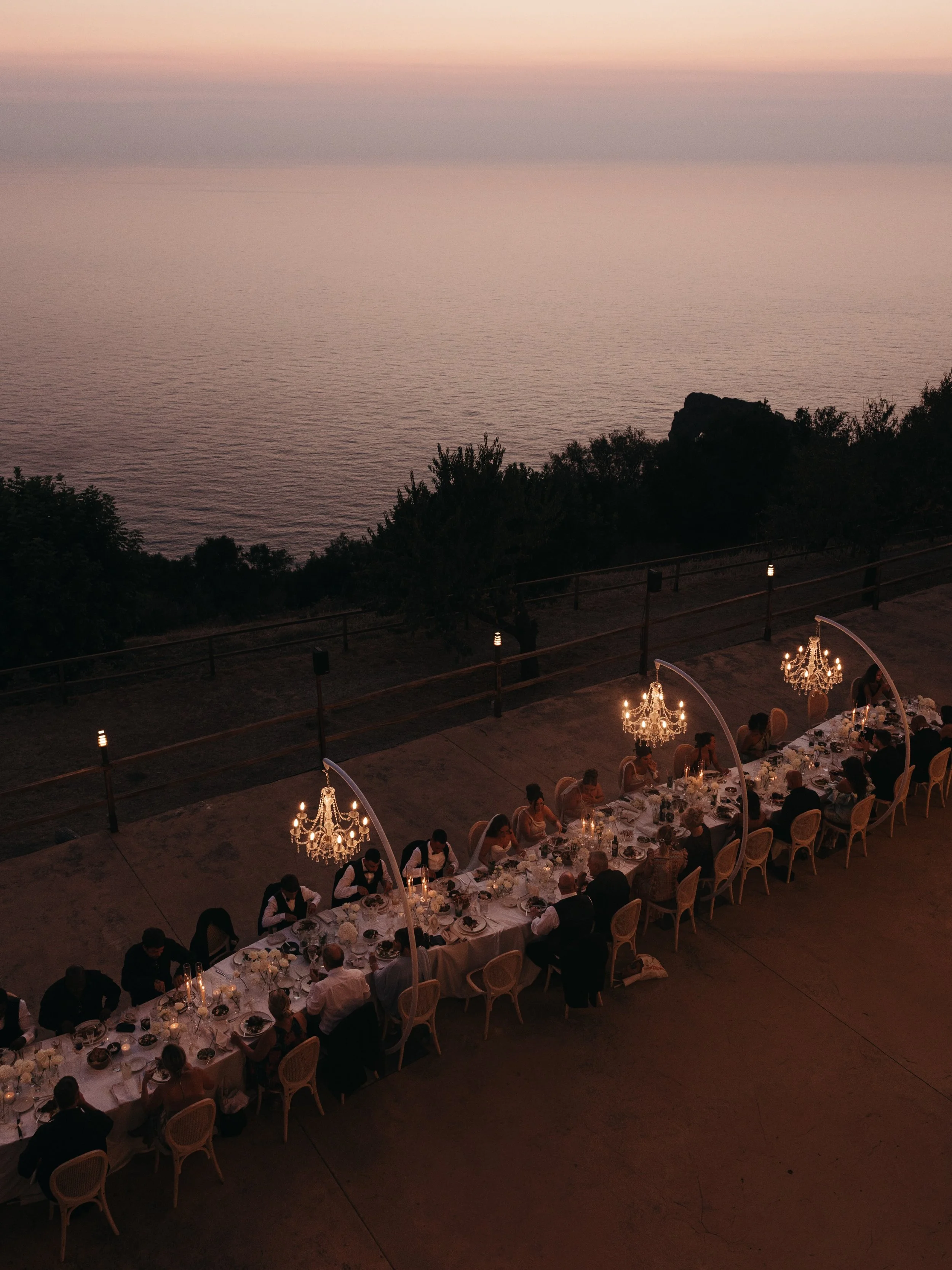 As the sun sets over Mallorca, dinner unfolds above the Mediterranean at Son Marroig. One long table, candlelight and chandeliers suspended beneath the open sky - an evening designed around the landscape, where the last light over the sea becomes par