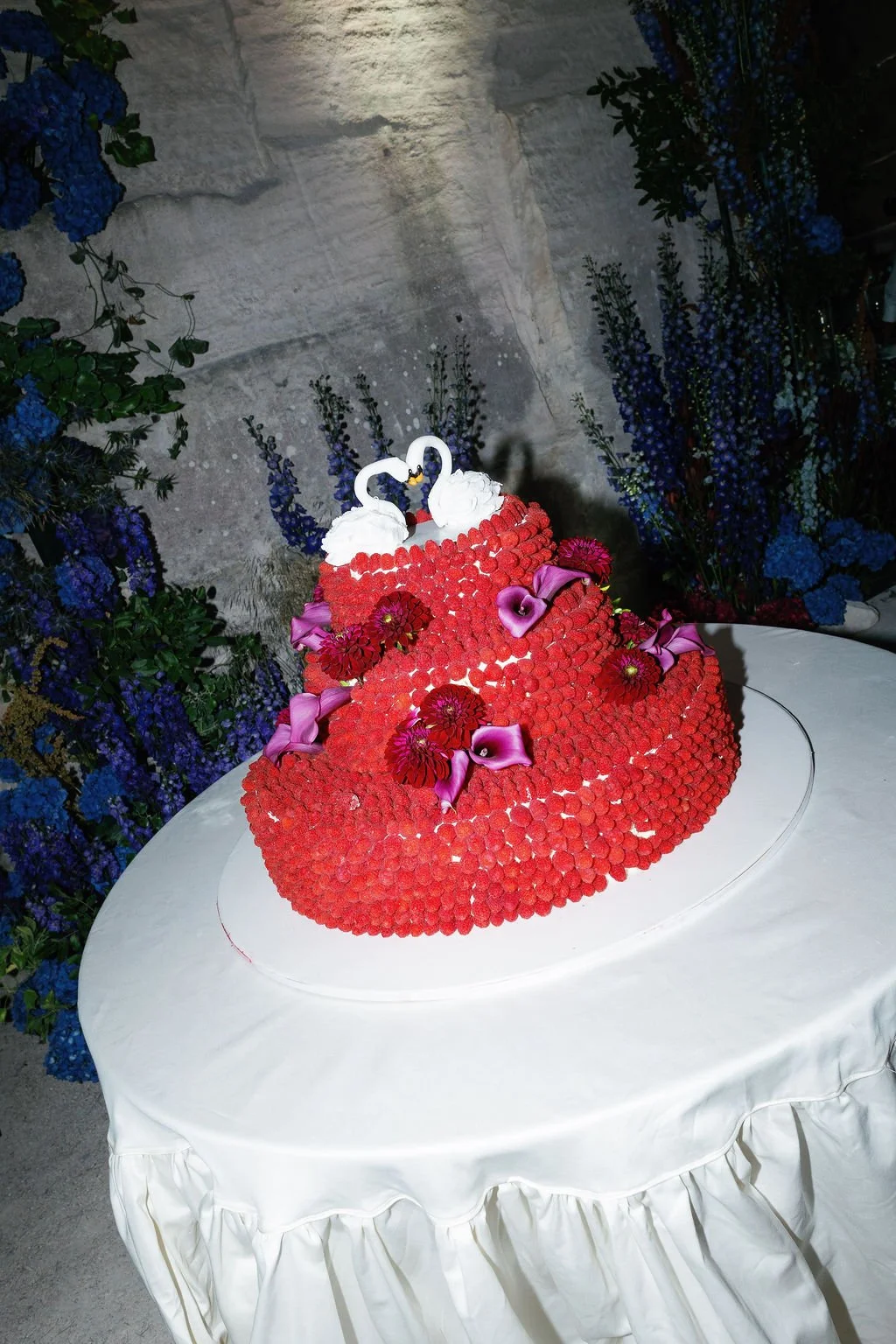 A heart-shaped cake decorated with red flowers and purple calla lilies, topped with two swan figurines and white whipped cream, displayed on a white draped table surrounded by blue and purple flowers.