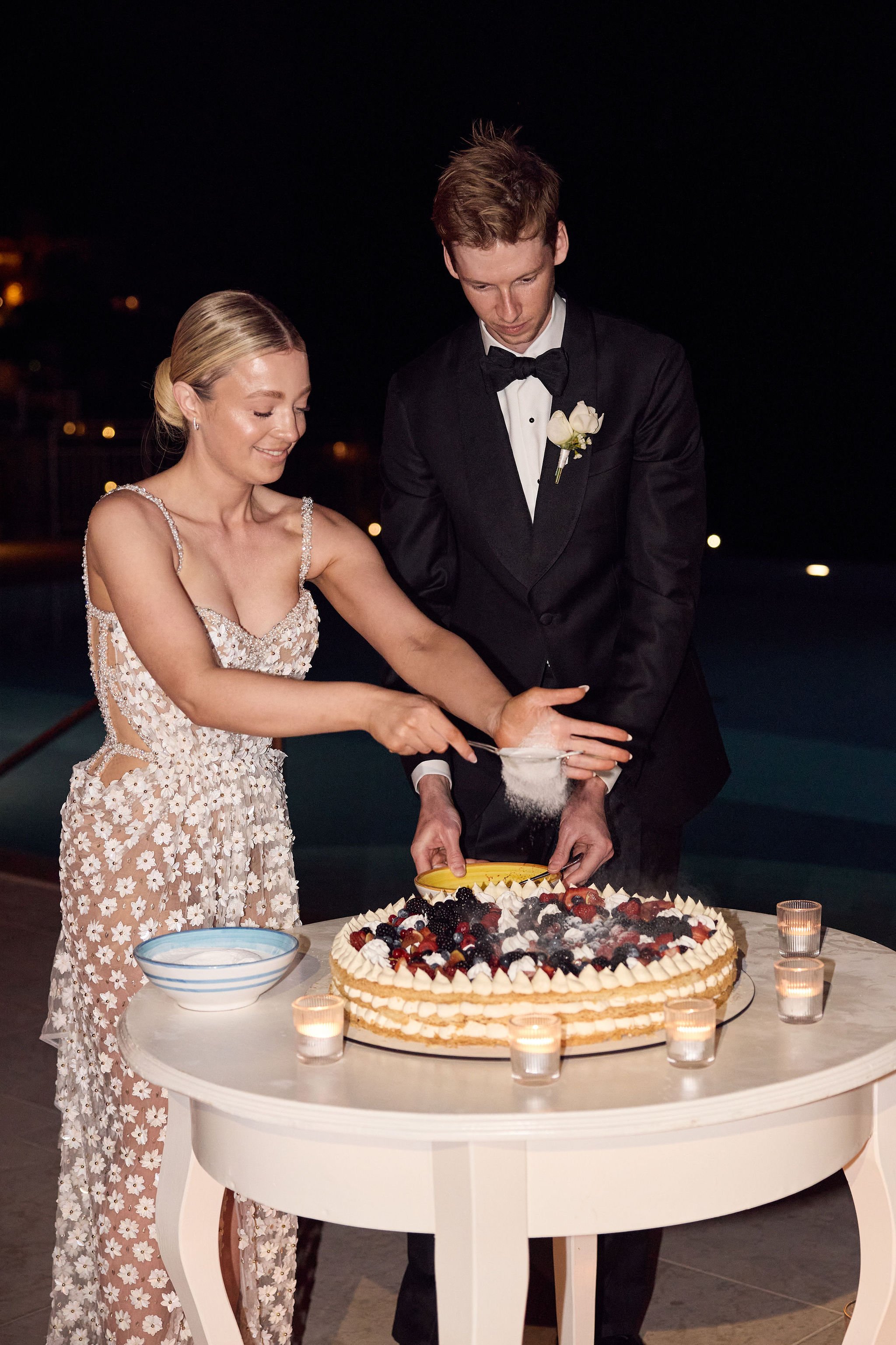 A bride and groom cutting their wedding cake at night, with candles on the table and a pool in the background.