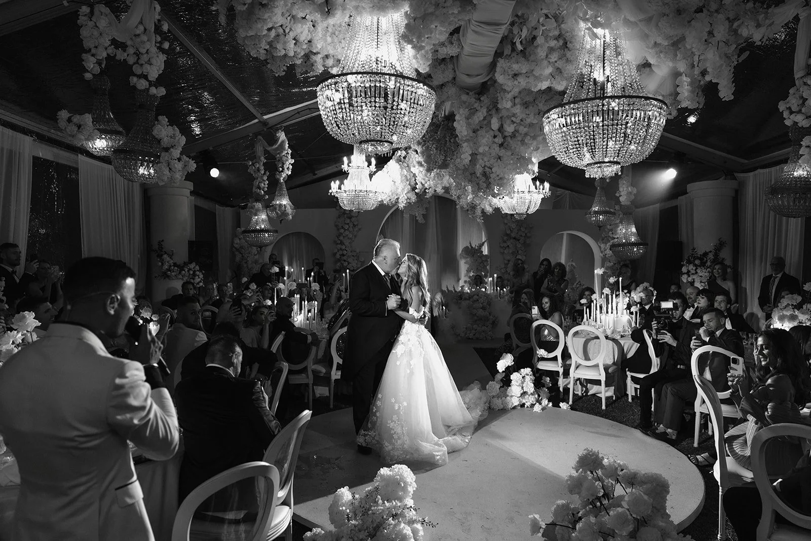 A wedding couple dancing in the center of a decorated banquet hall with guests seated around, chandeliers hanging from the ceiling, and floral arrangements.