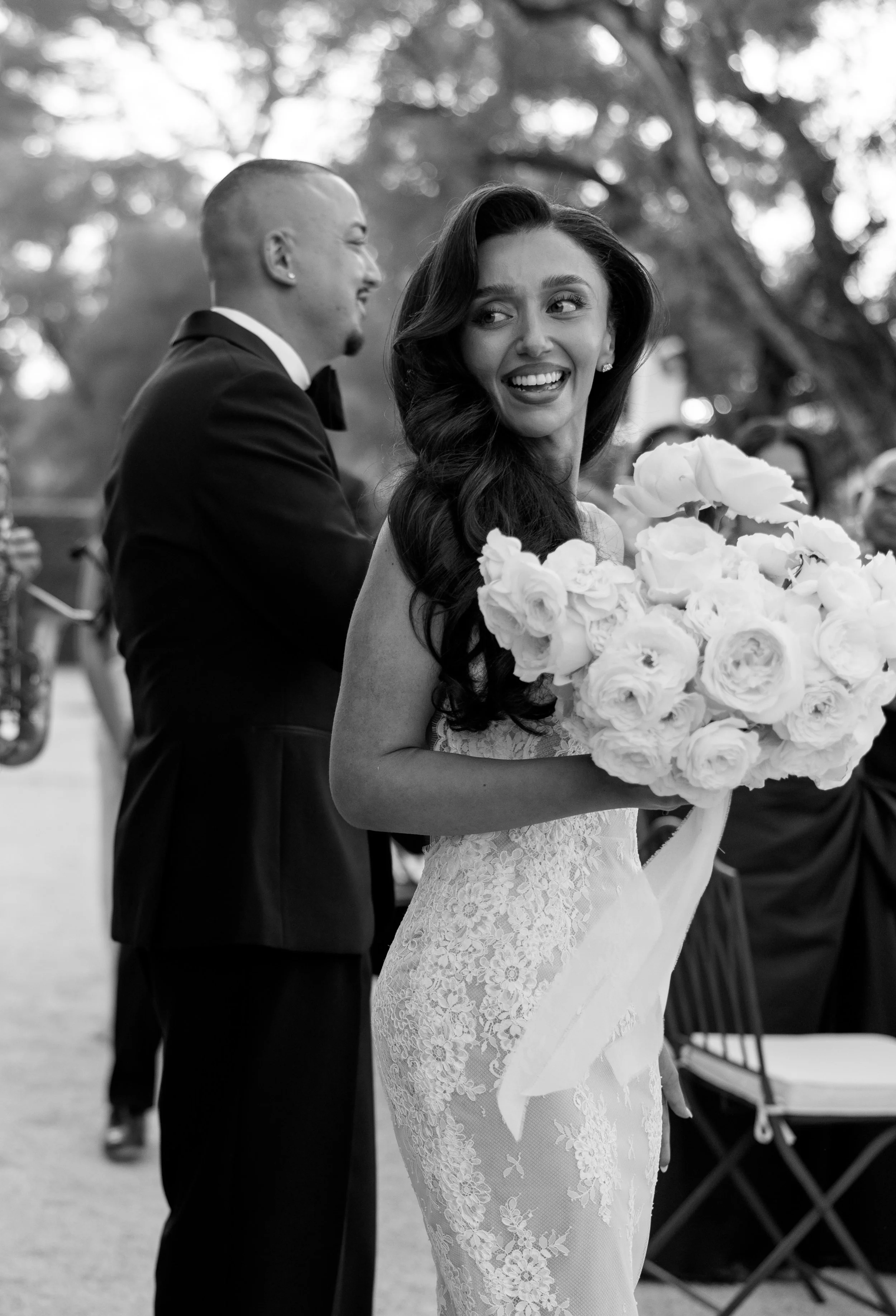 A black and white photo of a bride in a lace wedding dress holding a bouquet of flowers, smiling and looking to her right, with a man in a tuxedo standing behind her, at an outdoor wedding ceremony.