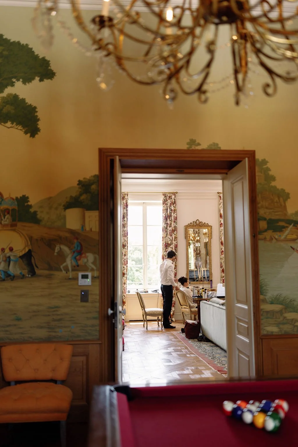 A view through a doorway into a well-lit room where a person is getting ready in front of a mirror, with a large window and floral curtains, in a room with elegant decor and wooden furniture.