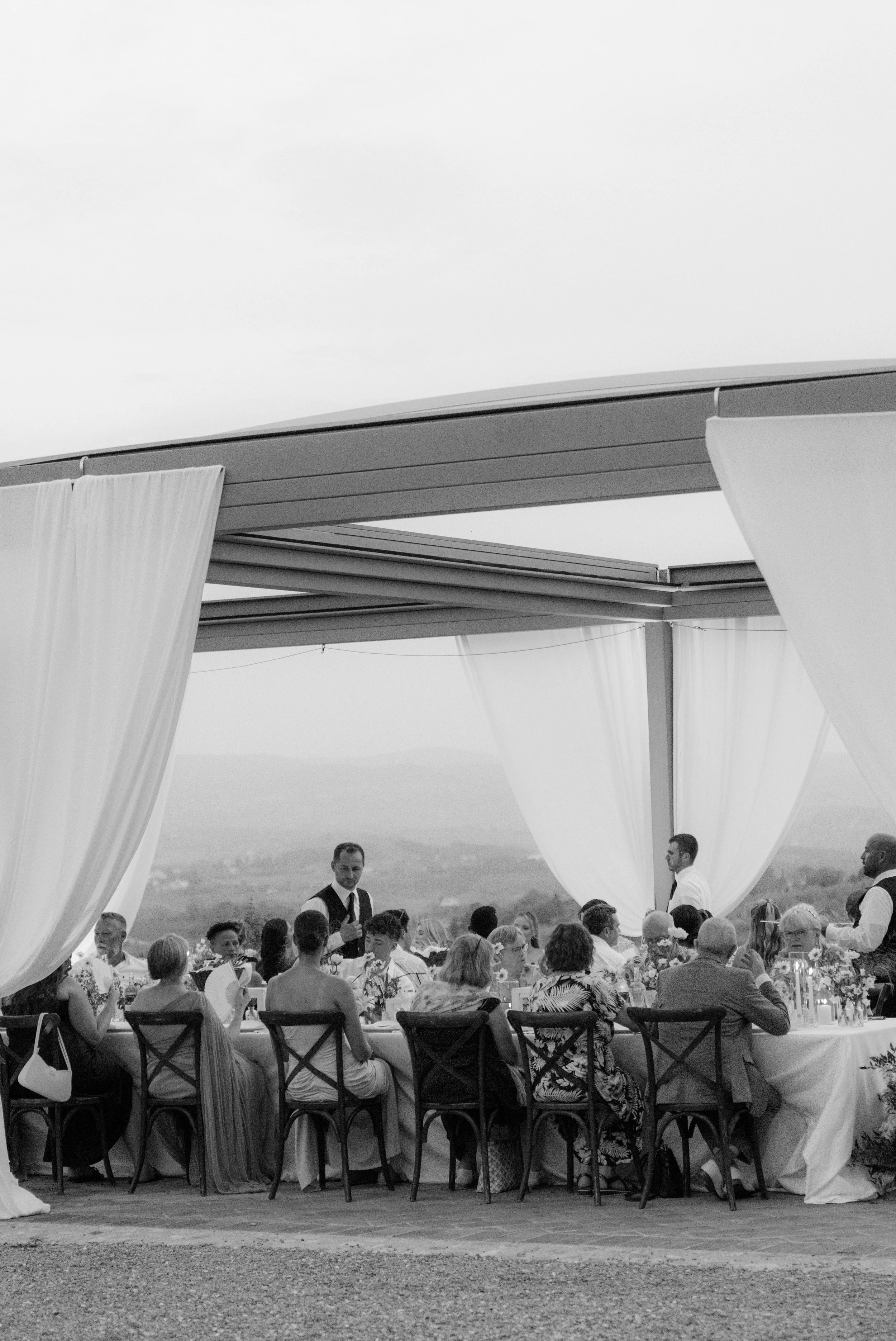 A black-and-white photo of a large outdoor gathering. People are seated at a long, decorated table with white tablecloths, surrounded by chairs. Two servers or waitstaff are standing and attending to the guests. The gathering is under a large canopy 