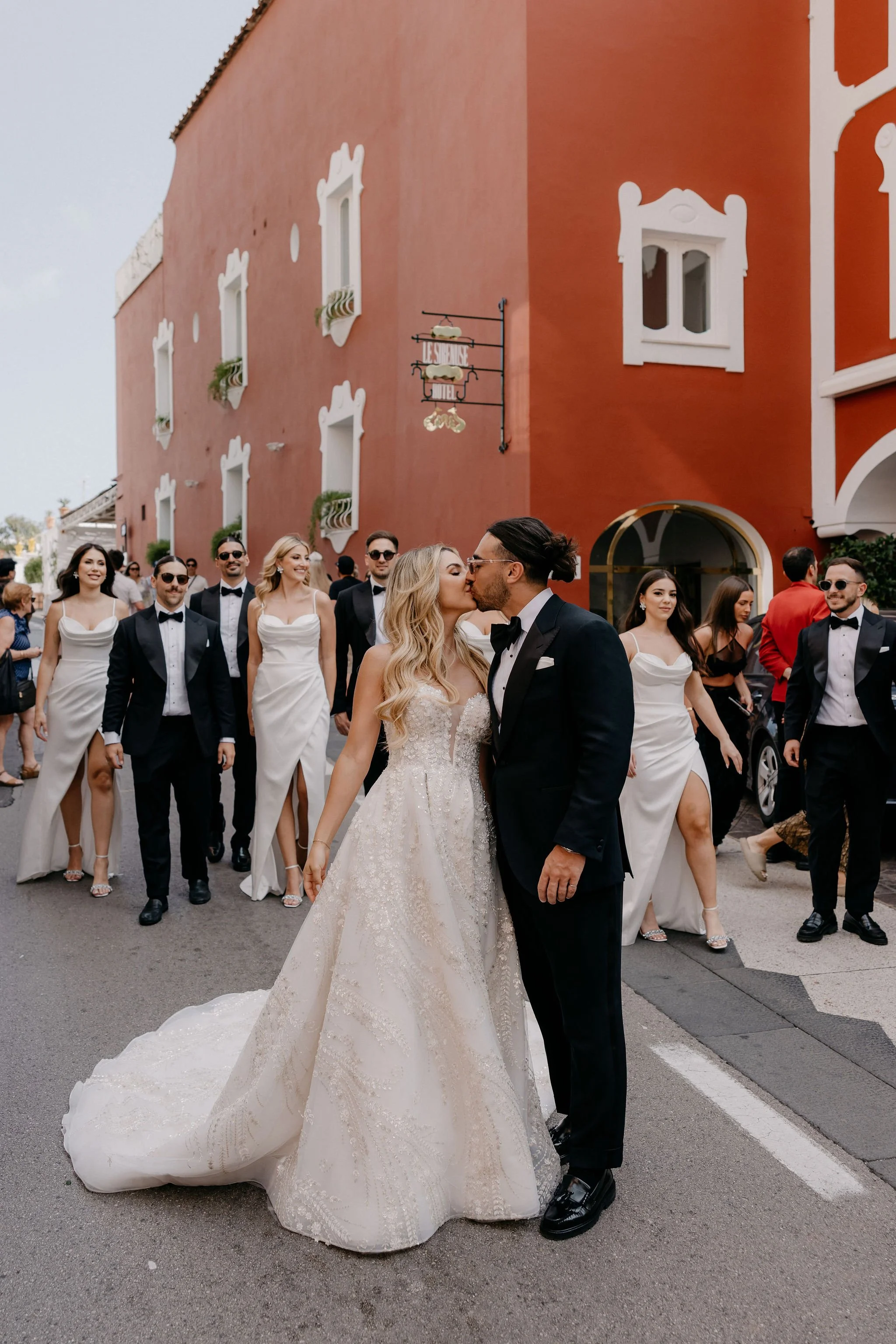Bride and groom kissing on a city street in front of a red building, surrounded by their wedding party.