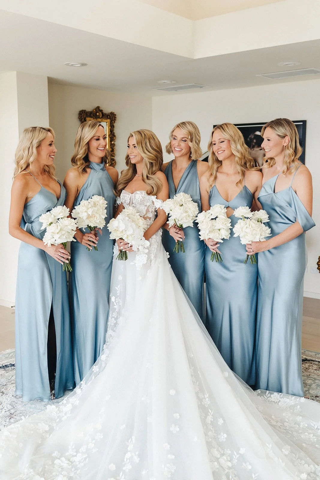 Bride and six bridesmaids dressed in matching blue gowns holding white bouquets, standing together indoors.