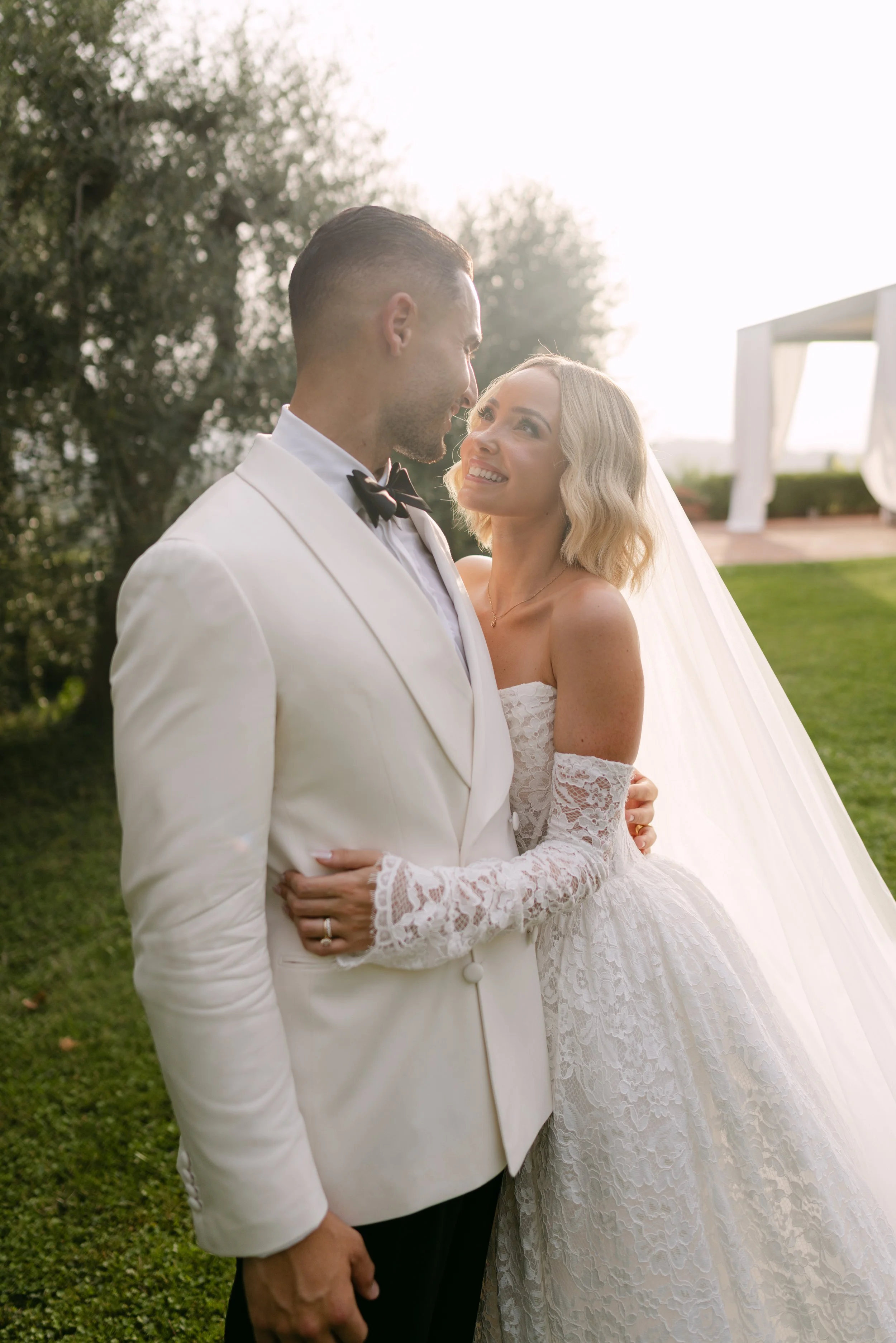 A bride and groom stand close together outdoors during sunset, gazing into each other's eyes. The groom wears a white tuxedo with a black bow tie, while the bride wears a lace wedding gown with a veil. Trees and a canopy are in the background.