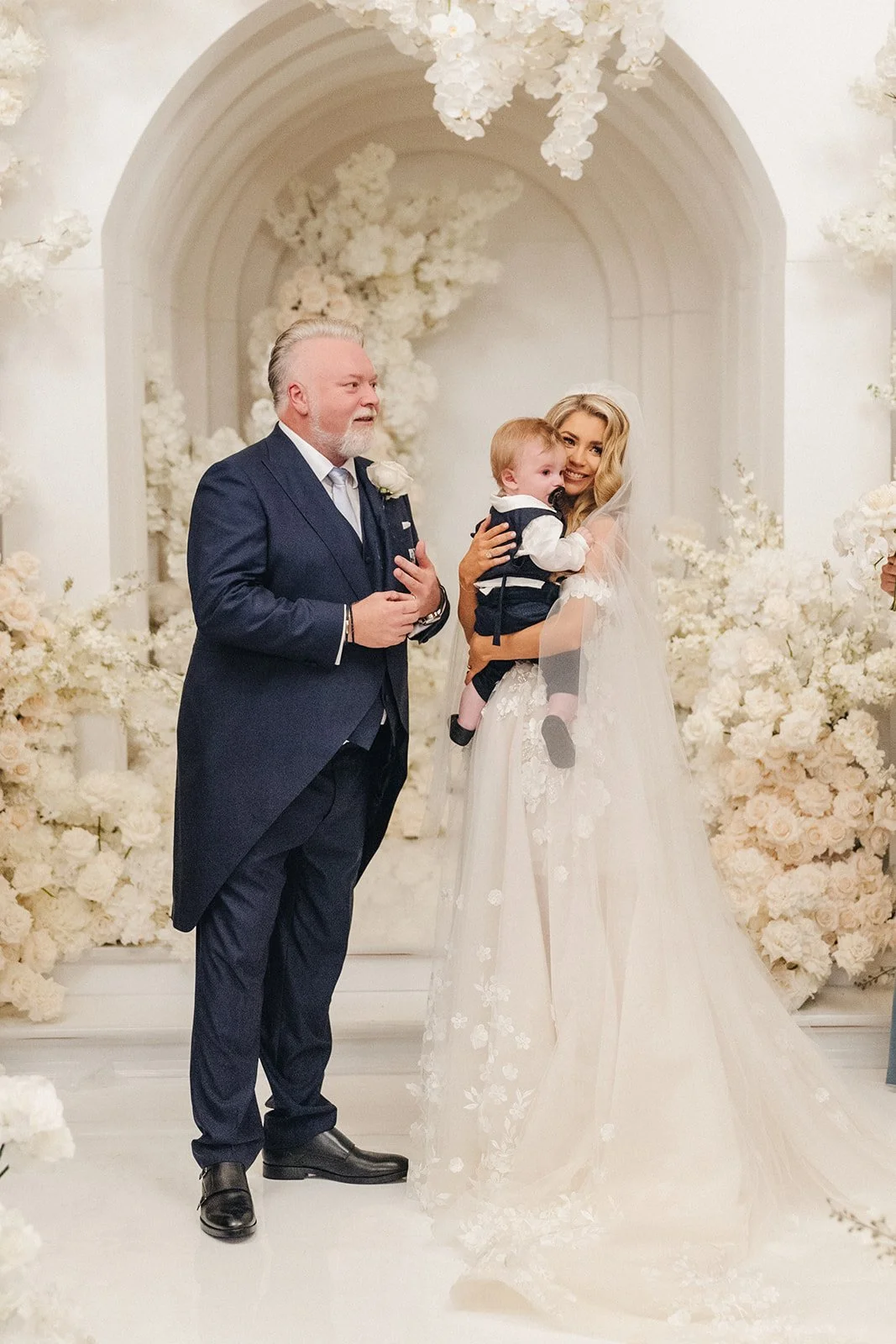 A bride holding a young boy dressed in a suit, standing next to an older man in formal attire in a wedding setting with white floral decorations.