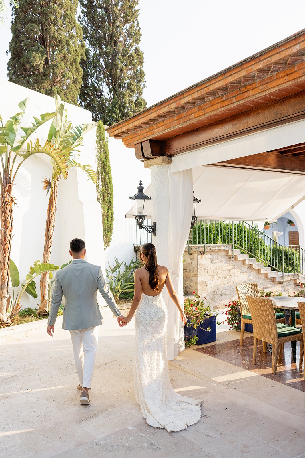 Couple in wedding attire walking hand-in-hand outdoors near white walls, lush plants, and outdoor seating area with chairs and tables.