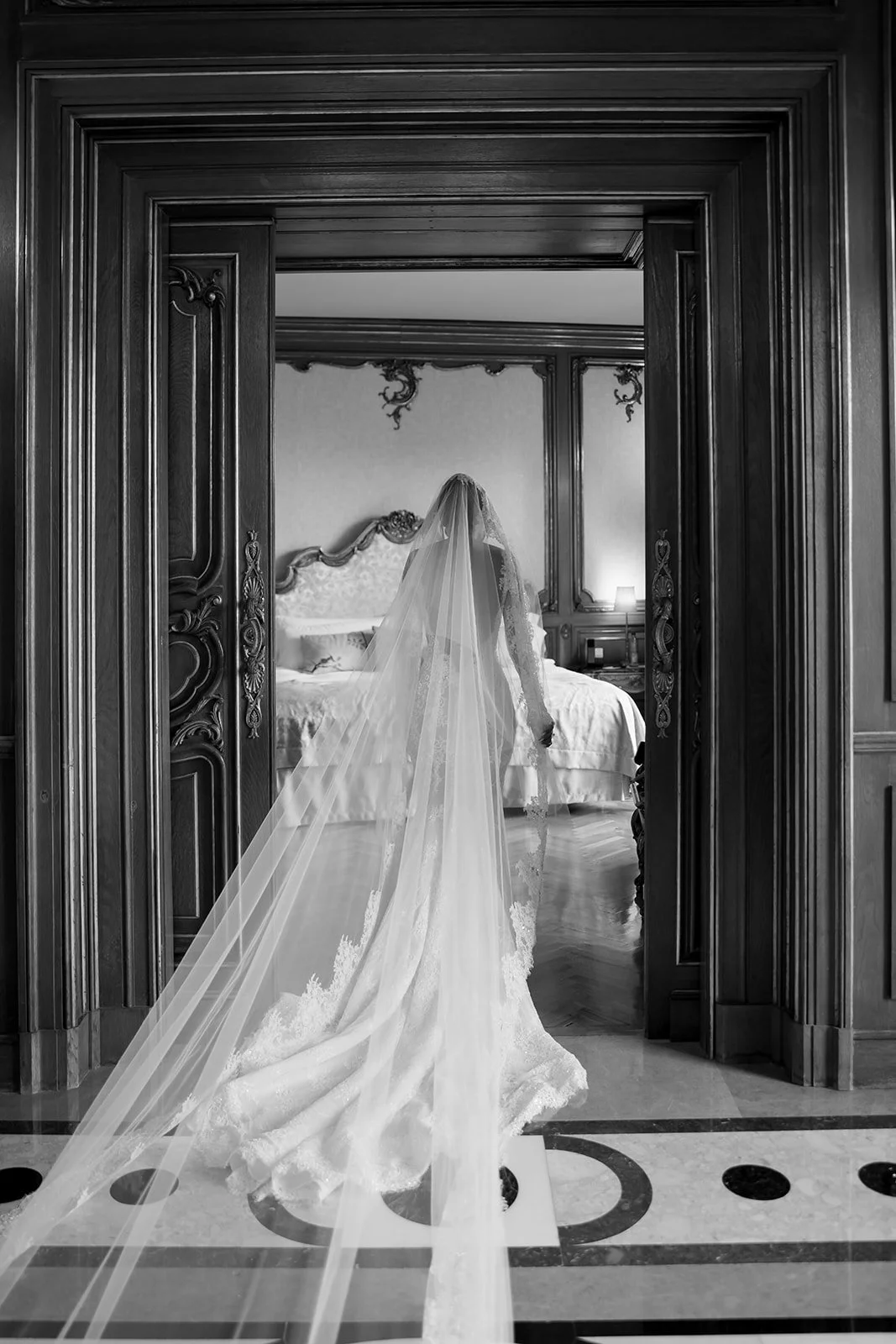 A bride in a wedding dress and veil stands in a lavishly decorated room, viewed through ornate wooden doorway.