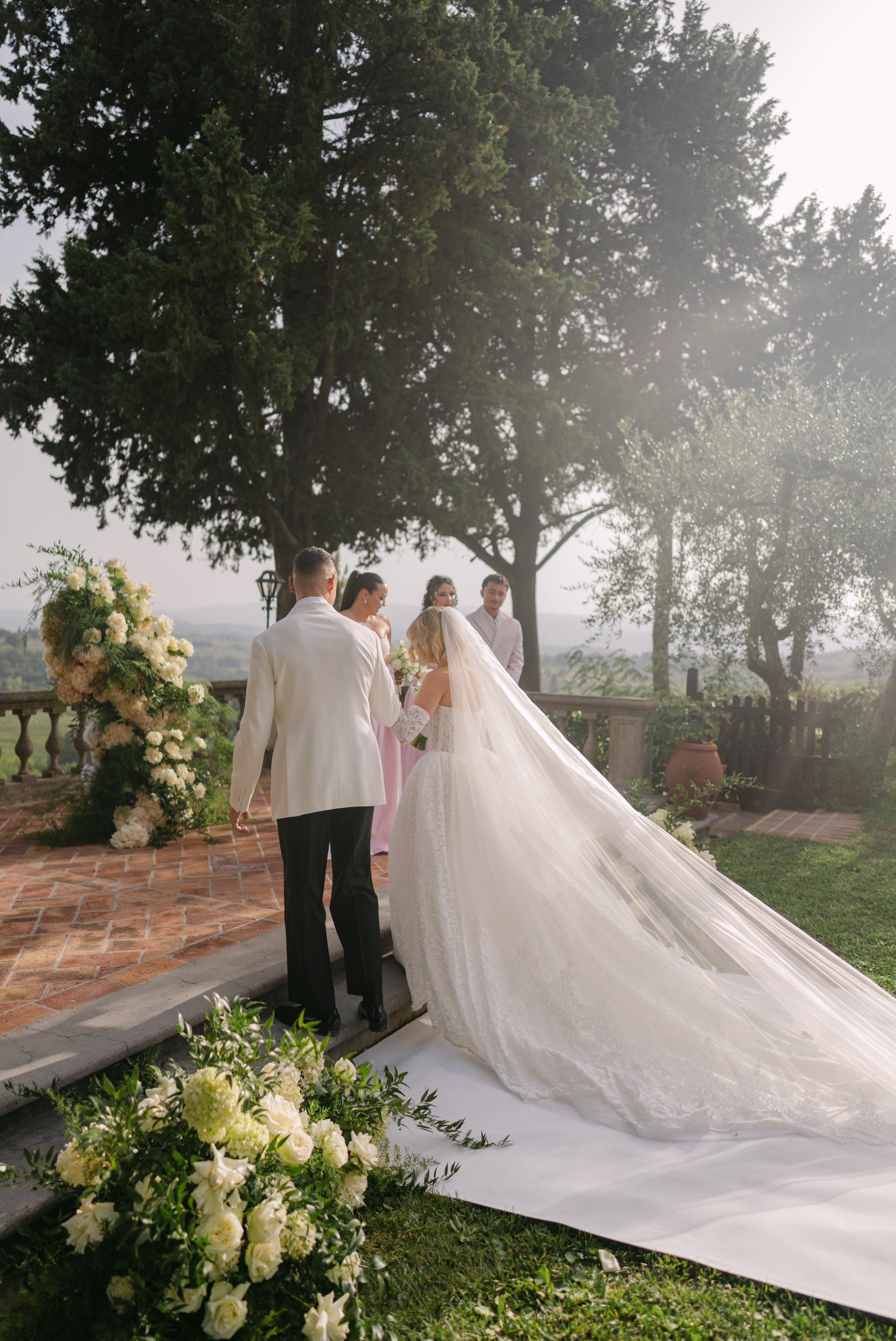 A wedding ceremony taking place outdoors with a bride in a white gown and long train, a groom in a white tuxedo, and three bridesmaids standing by a decorated altar with white flowers and greenery. Large trees and a scenic landscape are in the backgr