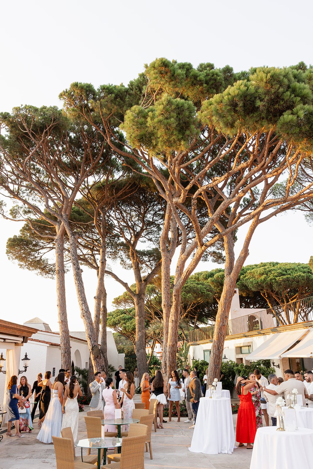 People gathering in an outdoor courtyard decorated for a celebration with large trees and white buildings in the background.