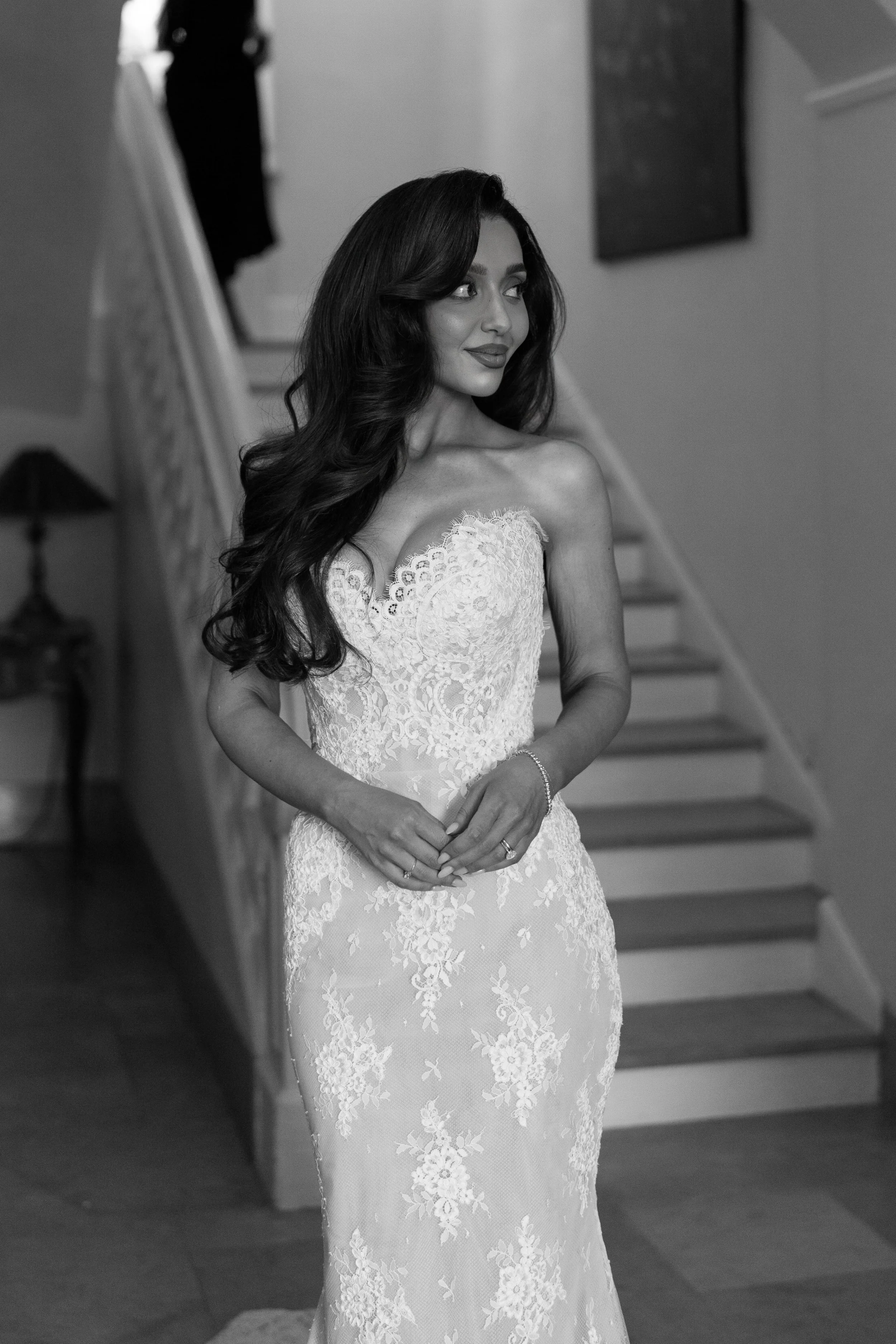 A woman in a strapless lace wedding gown standing on a staircase, smiling and looking to the side.