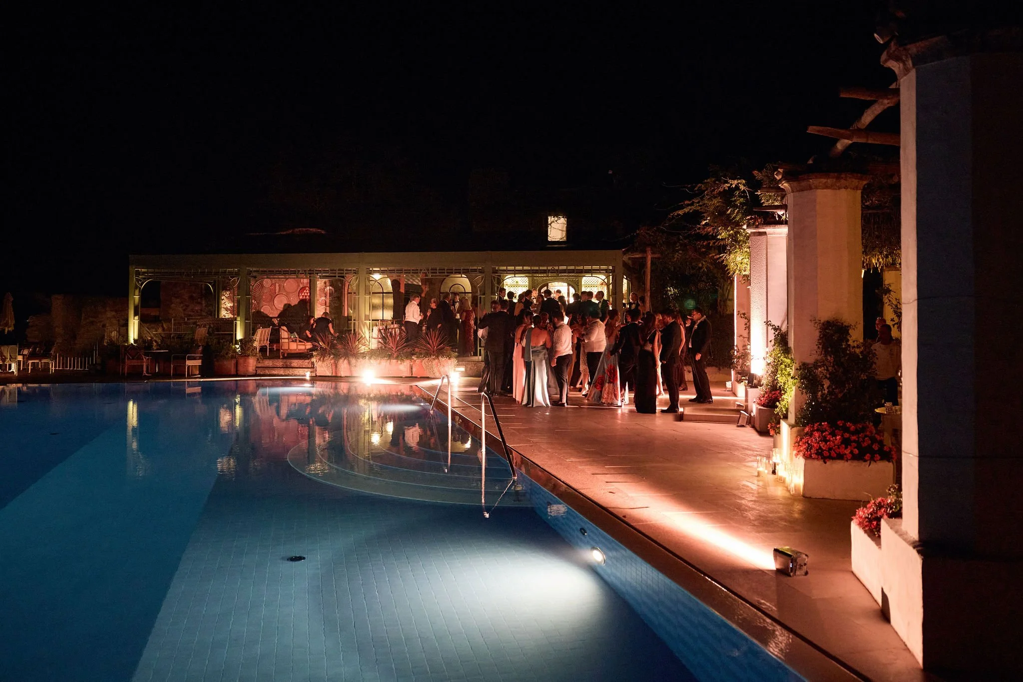 People gather in formal attire at an outdoor nighttime poolside event with ambient lighting and decorative plants.