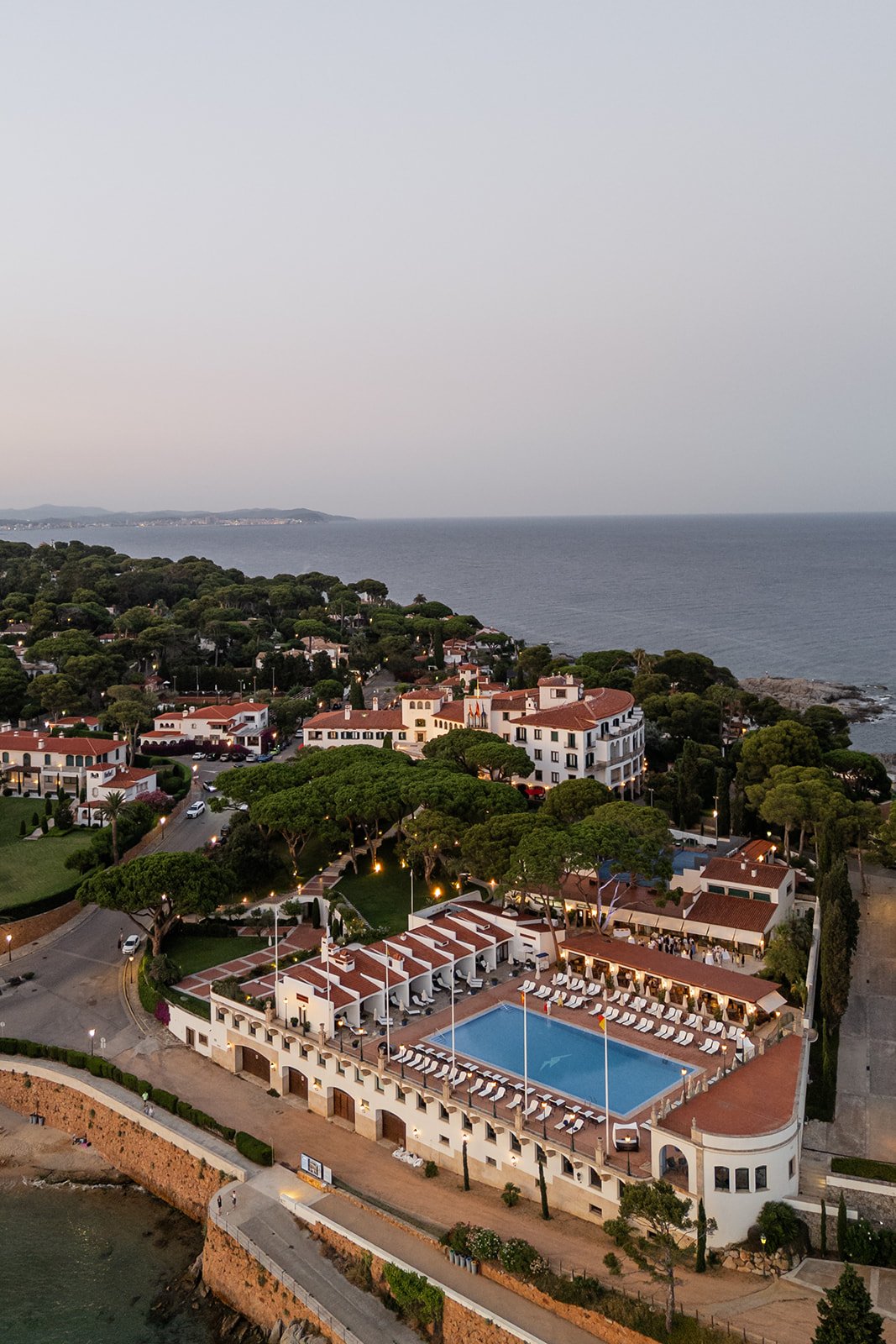 Aerial view of a coastal resort town with a large swimming pool, surrounded by white buildings with red tile roofs, lush green trees, and a rocky shoreline in the background.
