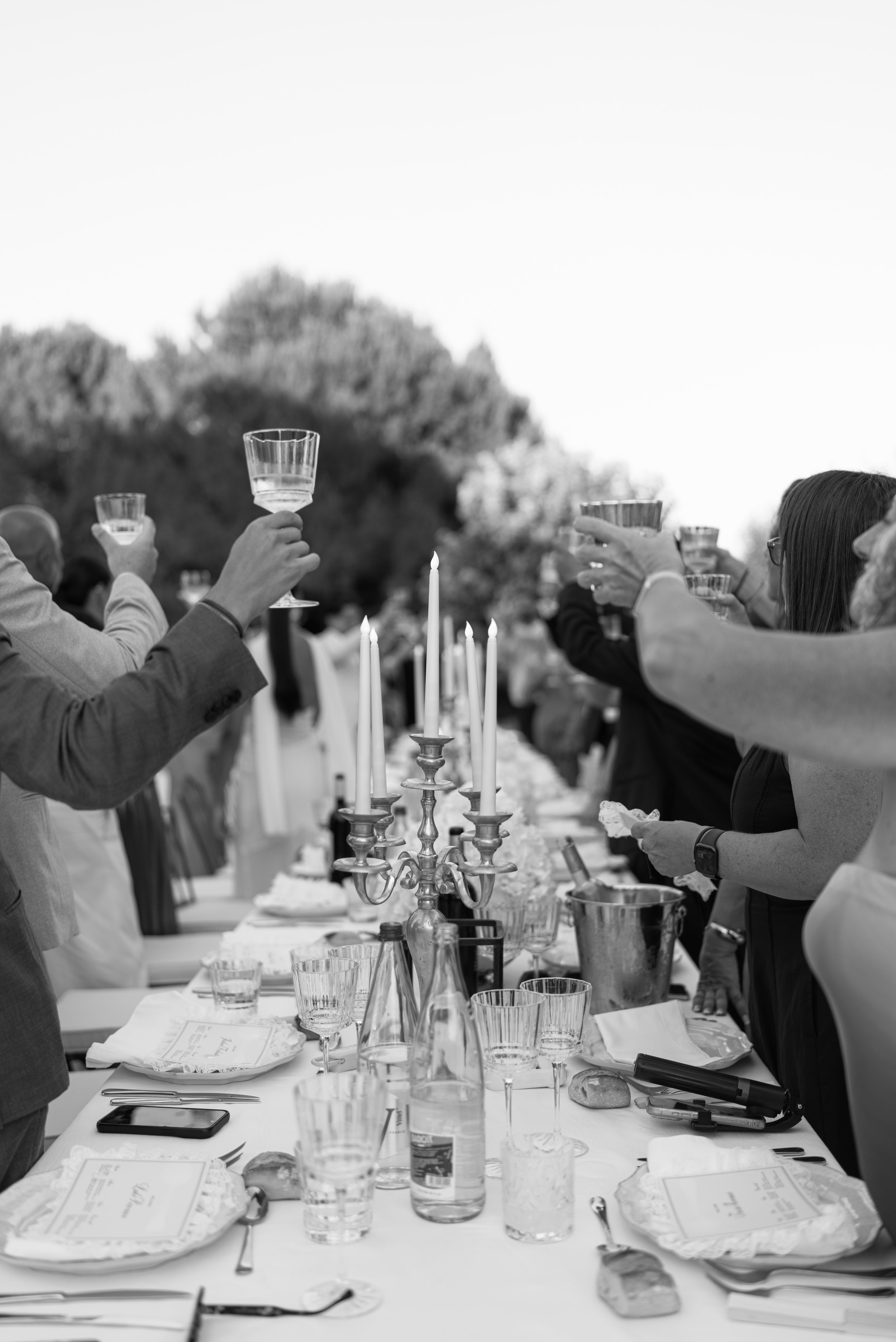 People raising glasses during a dinner party outdoors, with a long table set with plates, glasses, and a candelabra, in black and white.
