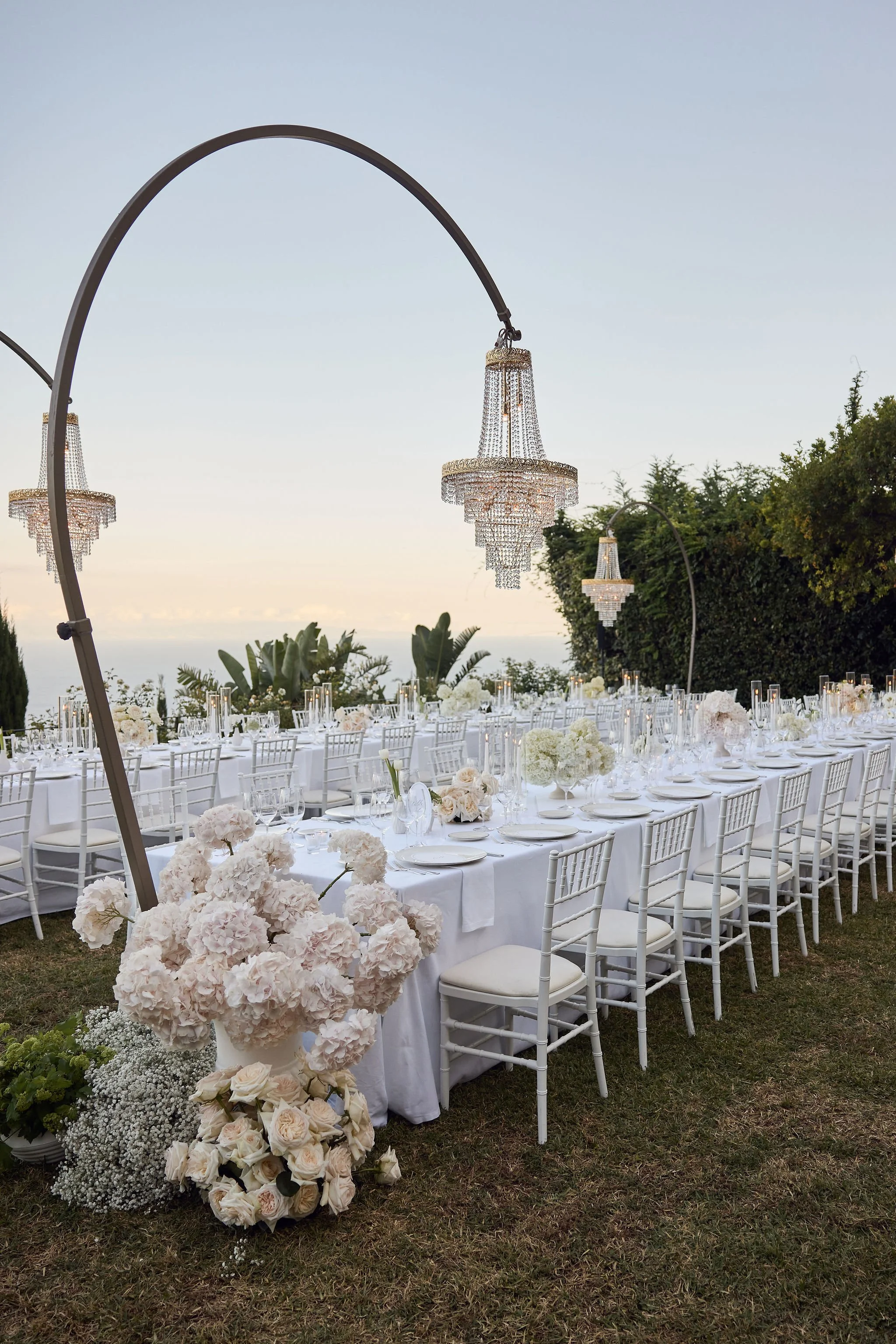 Elegant outdoor wedding reception setup with long white tables, white chairs, floral centerpieces, and crystal chandeliers hanging overhead, set against a scenic backdrop of trees and sky at sunset.
