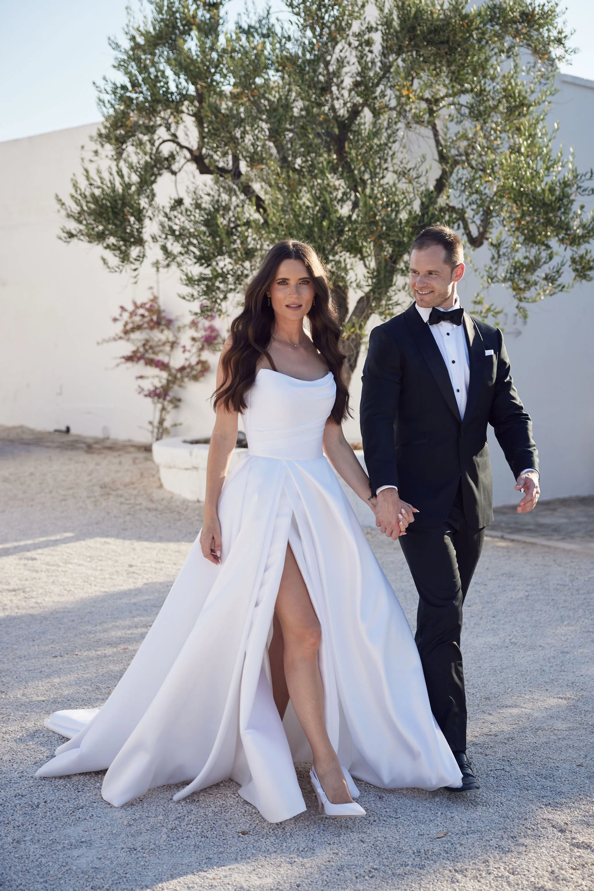 A newly married couple walking outdoors on a sunny day. The woman wears a white wedding gown with a high slit and white heels, and the man wears a black tuxedo with a bow tie.