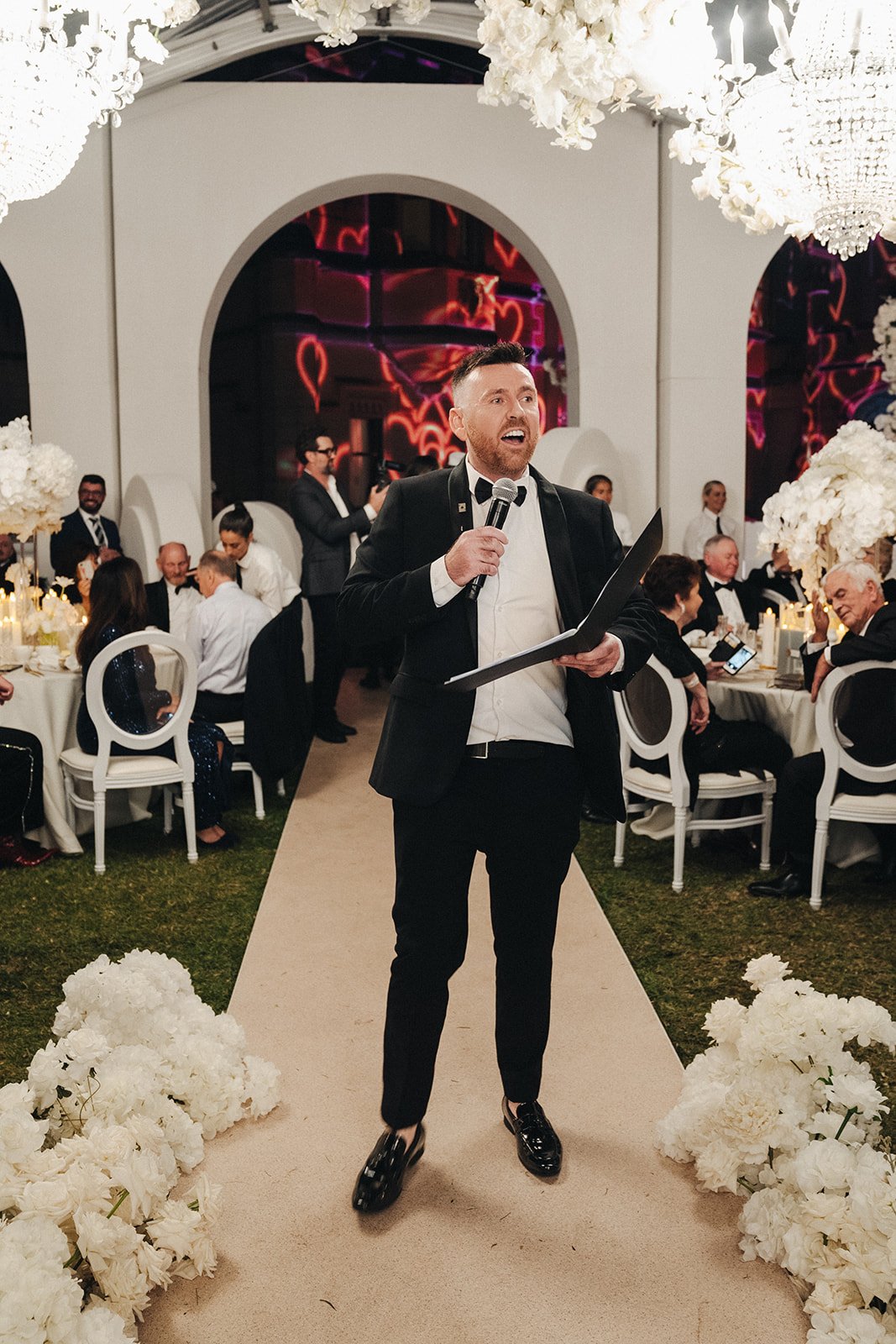 A man in formal attire, holding a microphone and a folder, is standing on a beige aisle at an elegant event with decorated tables and seated guests. The setting includes large white floral arrangements and chandeliers, with red neon heart signs in th