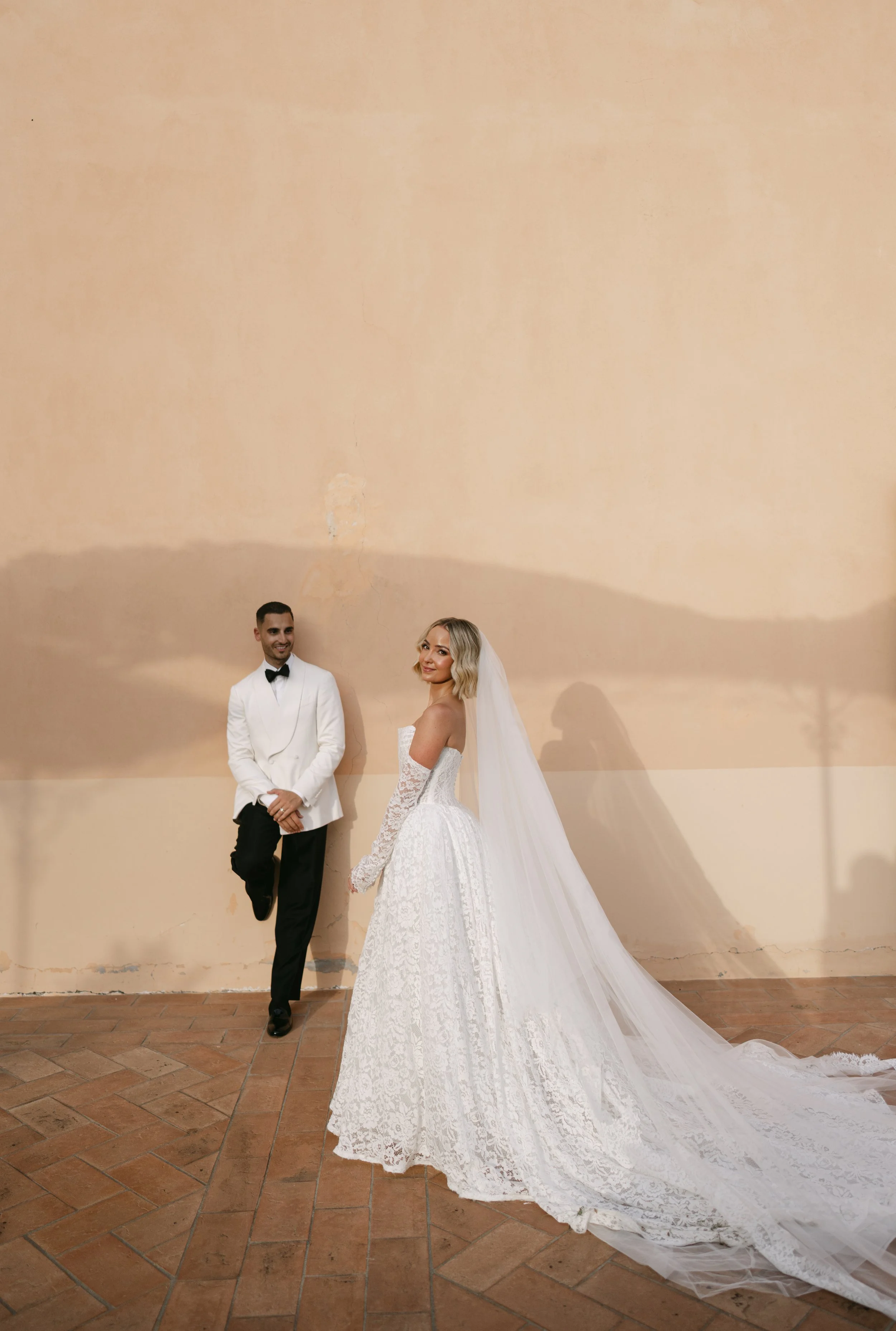 A bride and groom stand against a beige wall, holding hands. The groom is dressed in a white tuxedo jacket with black bow tie and black pants. The bride wears a long white gown with lace details and a veil flowing down her back.