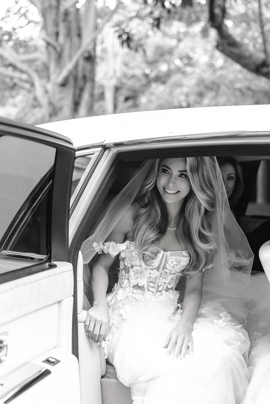 A bride with long wavy hair wearing a floral wedding dress and veil, smiling inside a vintage car.
